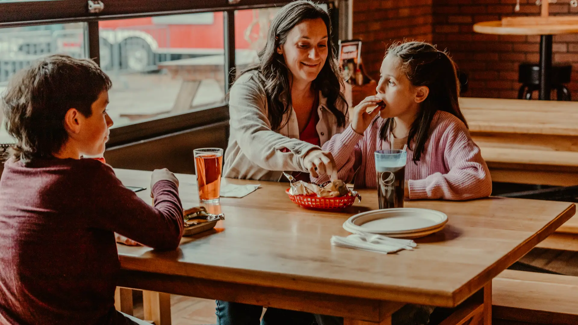 A mother and her kids enjoy a meal at big zs in front of the large glass garage doors.  Photo Credit to A. Kelly.
