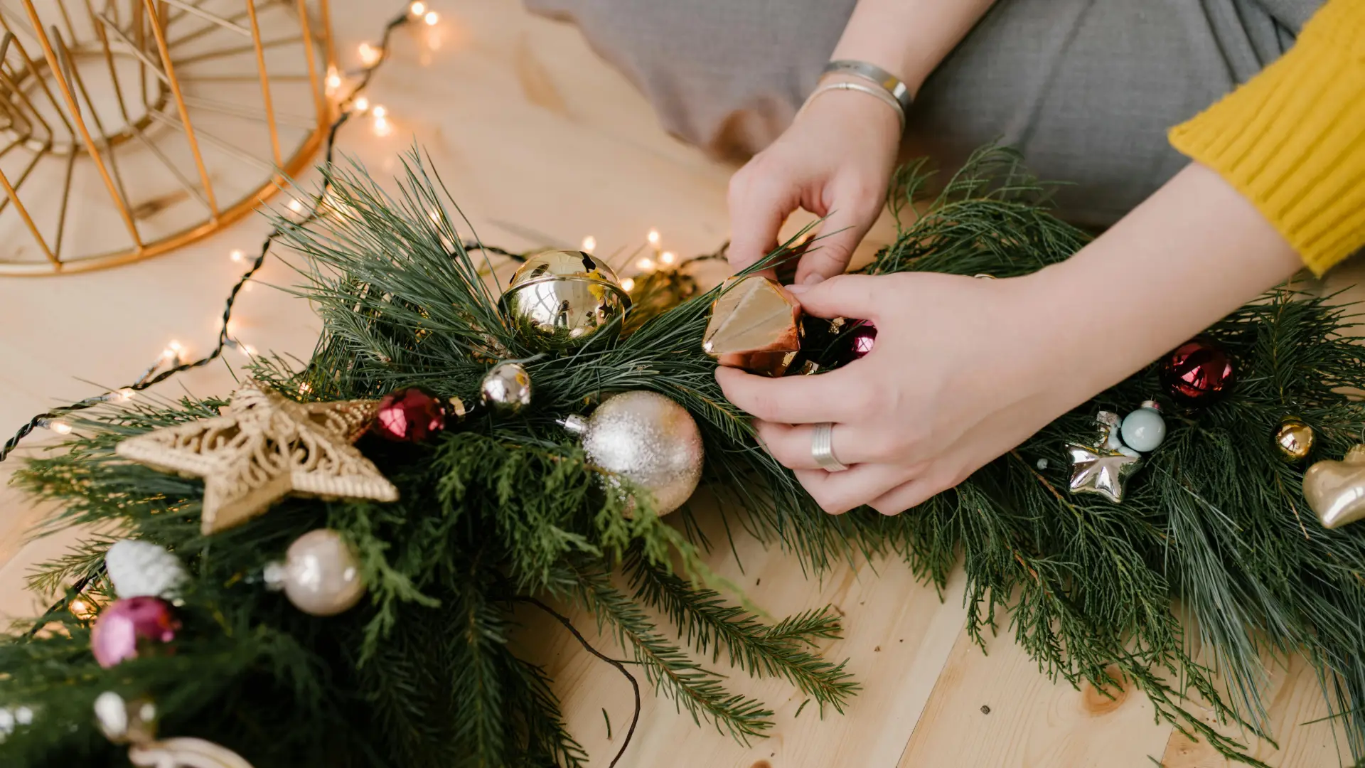 A close-up shot of a person arranging ornaments and pine sprigs to create a Christmas garland on a light wooden floor, surrounded by twinkling string lights.