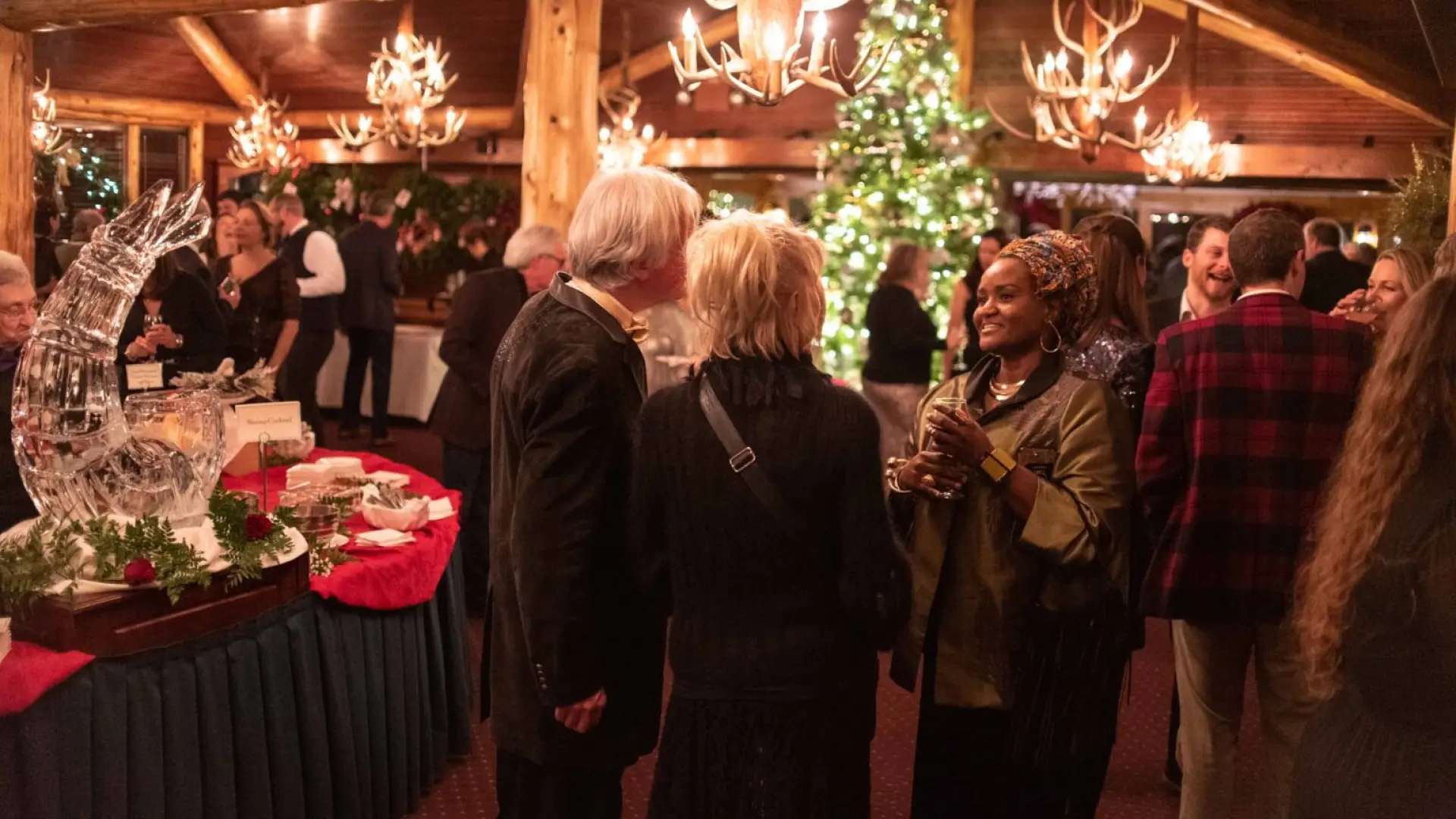 People mingle with drinks in hand amid buffet tables and Christmas trees and decor in an upscale lodge room