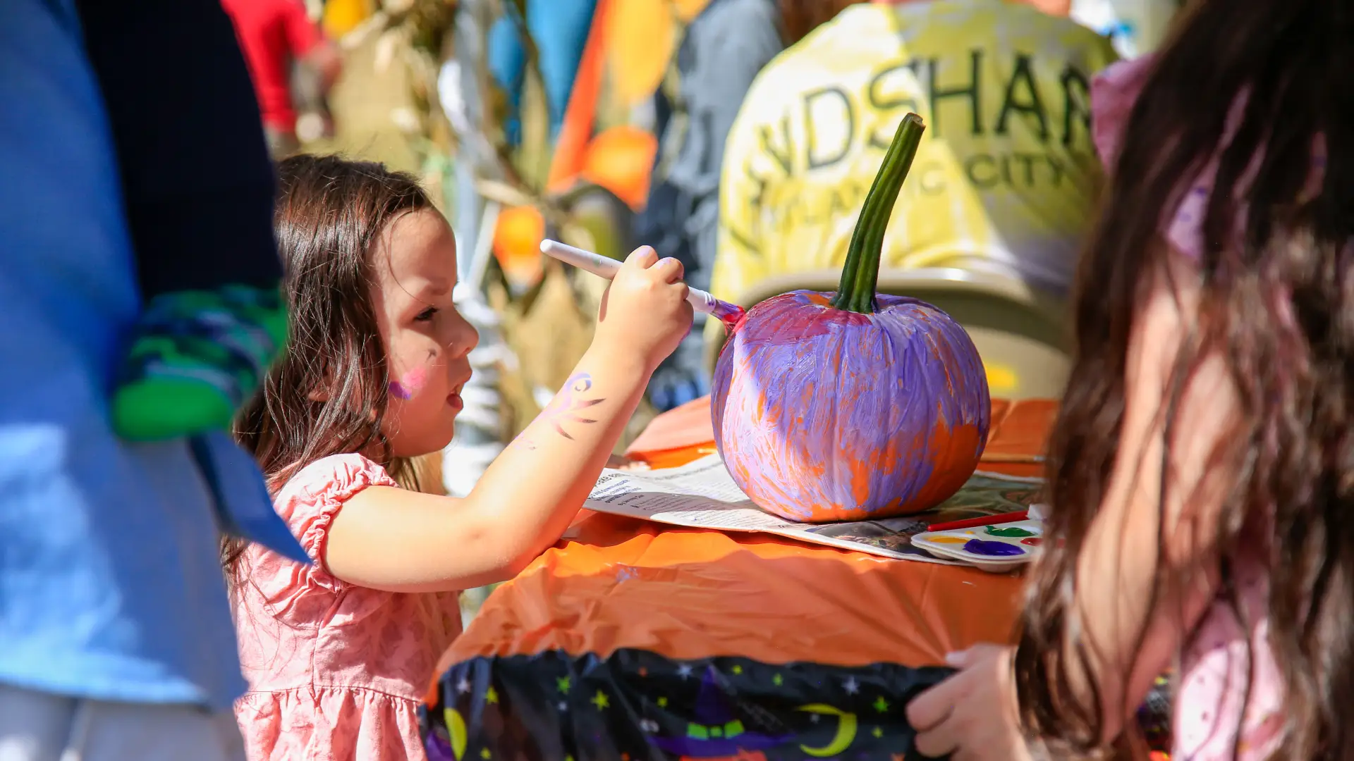 Young brunette girl in a pink dress paints a small pumpkin at a fall event