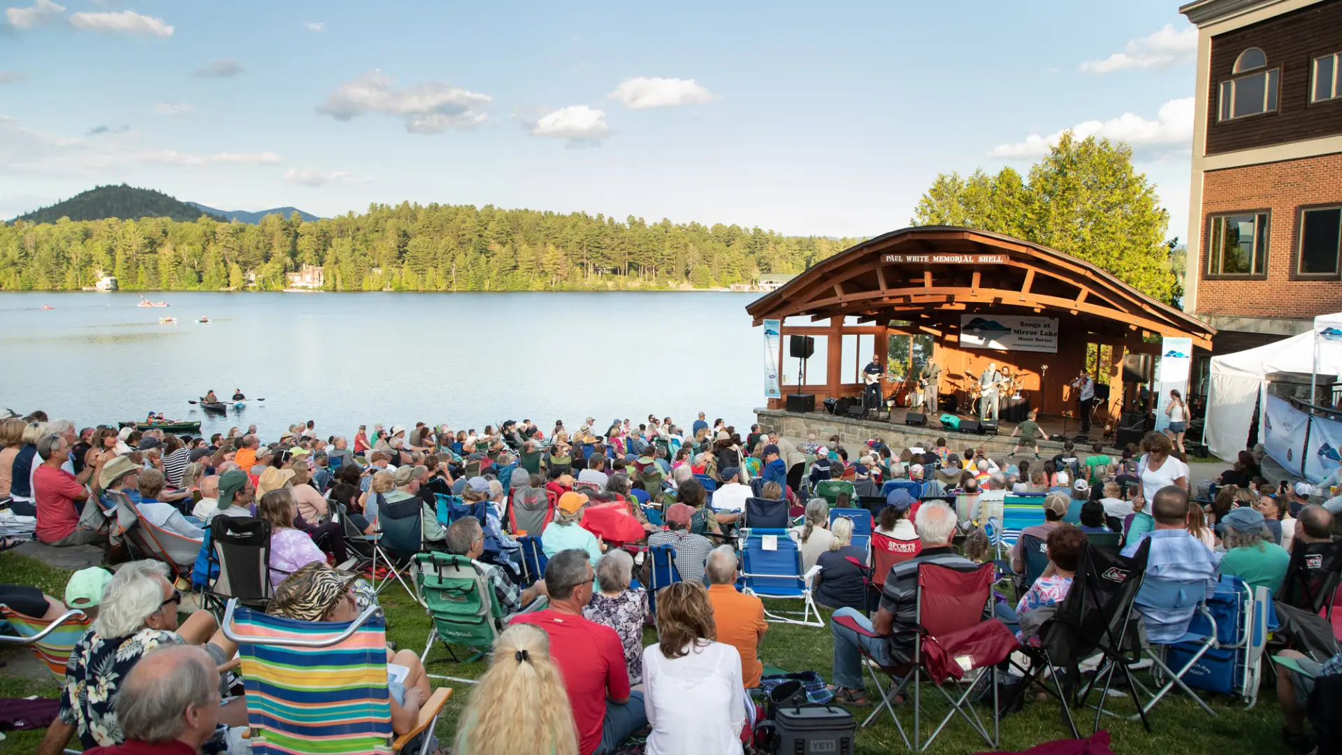 A lakeside park is filled with people in lawn chairs watching a concert in an outdoor bandshell.