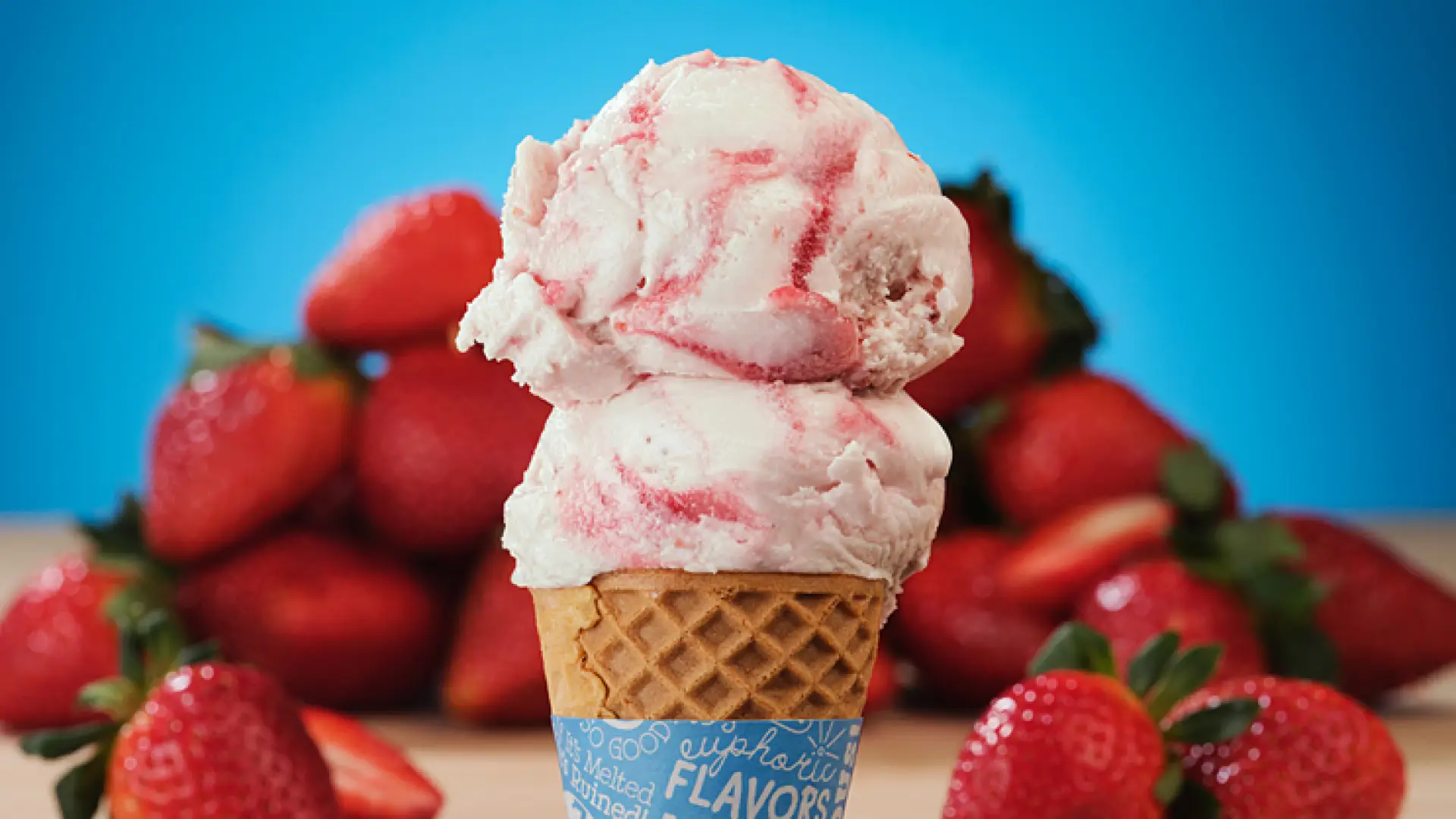 Double scoop of creamy strawberry ice cream in a waffle cone with blue background and ringed with red juicy strawberries