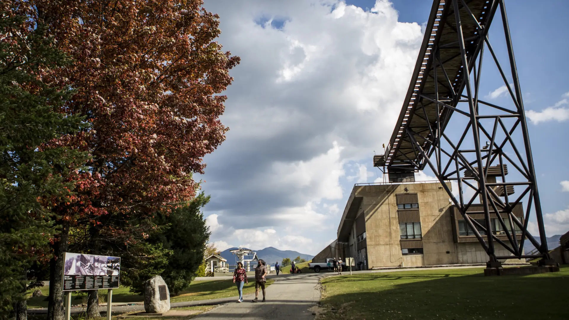 A couple walks on a path near a tree with red leaves and the towering Olympic ski jump.