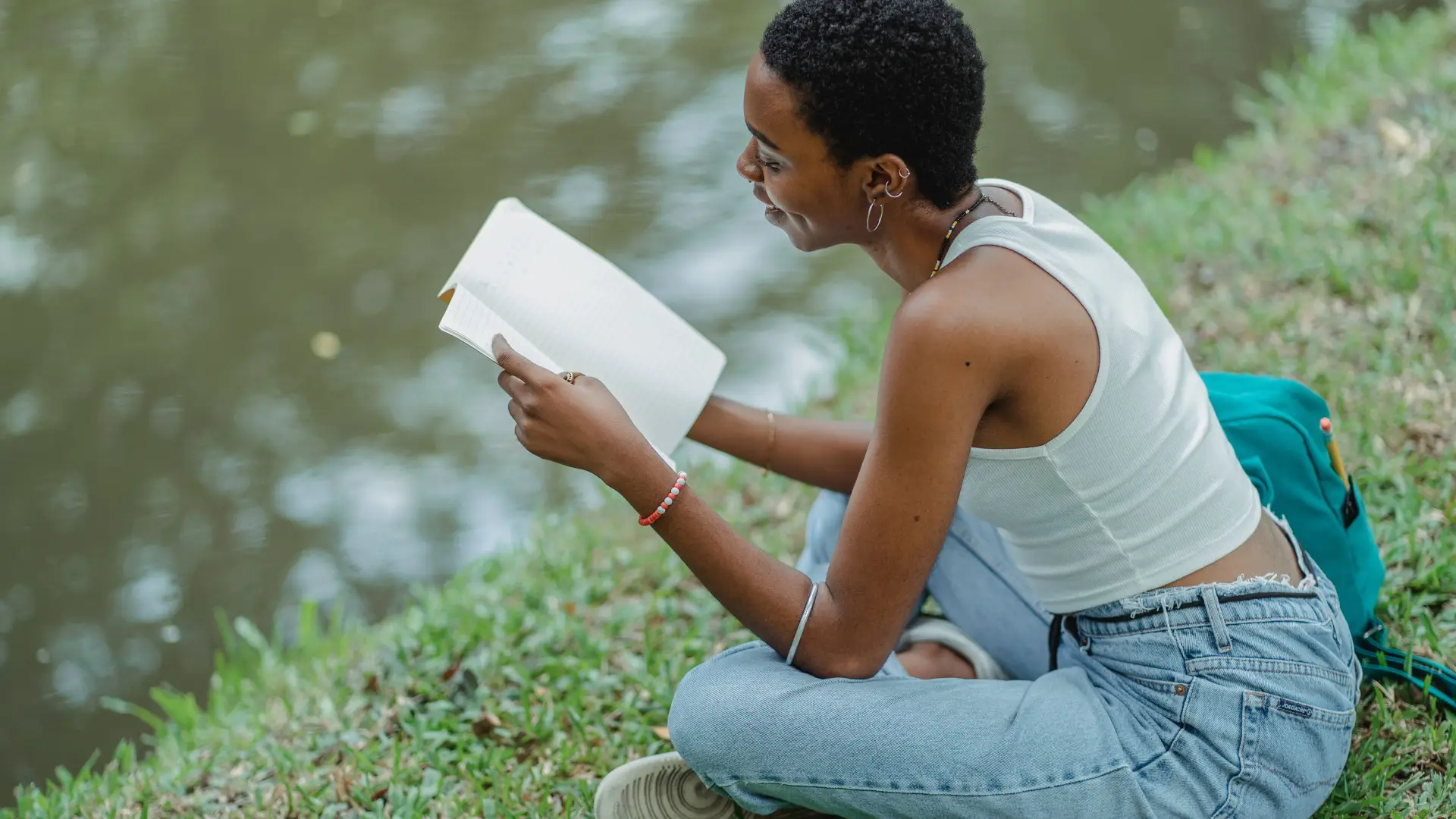 Young woman sits next to lake on grass reading a book wearing jeans and white tank top