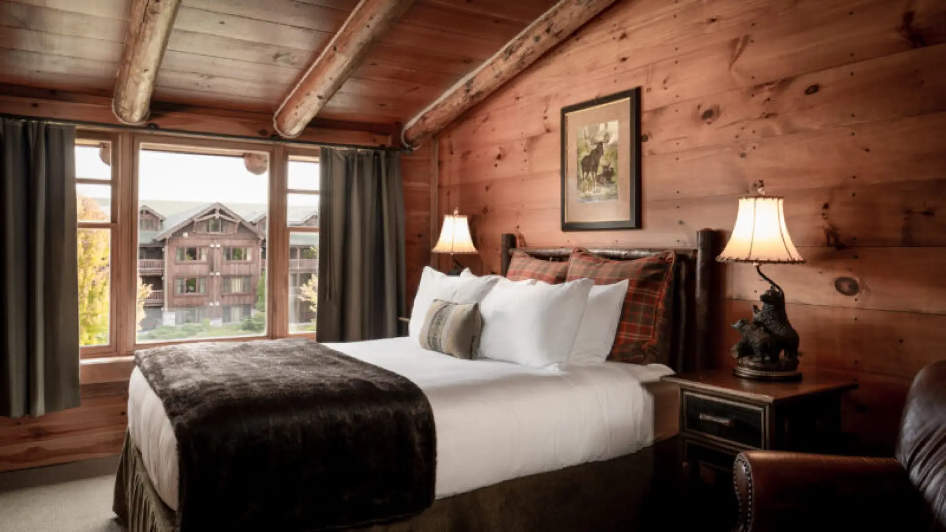 A luxurious bed with white sheets in knotty pine paneled room with thick beams and great views.