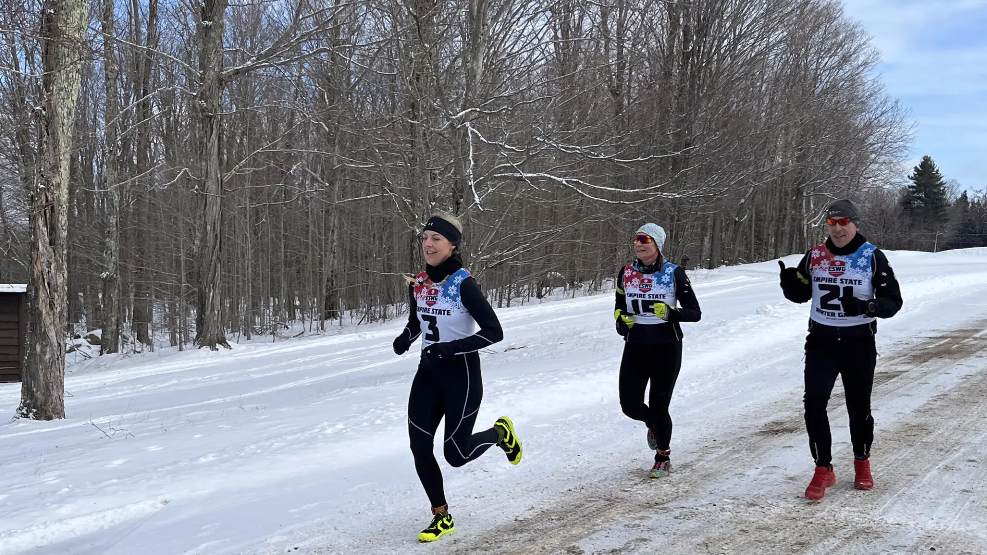 Three runners in winter gear race on a snowy, tree-lined path. The lead runner smiles, conveying determination and joy.