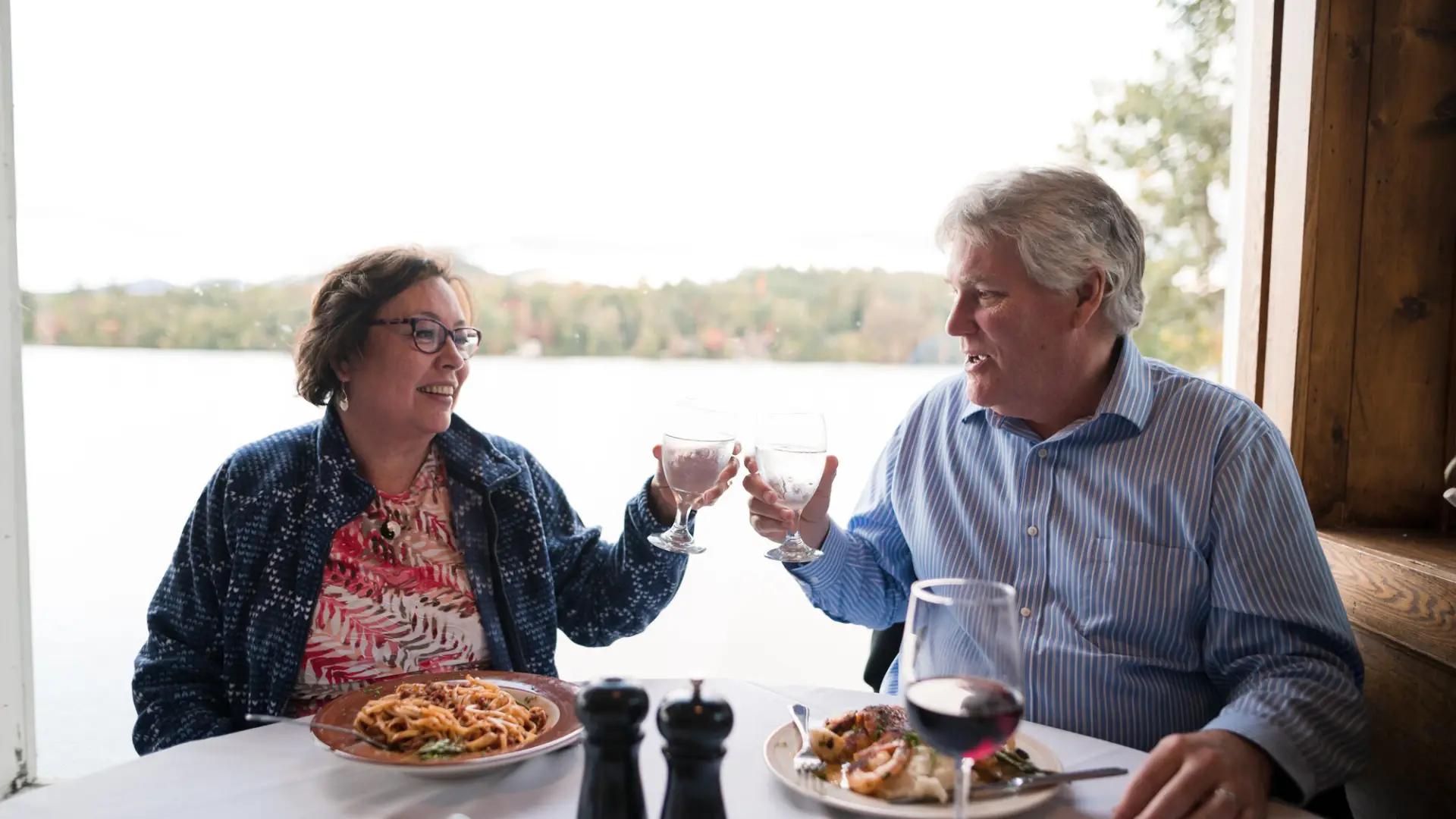 A couple (man and woman) clink drinks together at a restaurant table overlooking a lake and mountains.