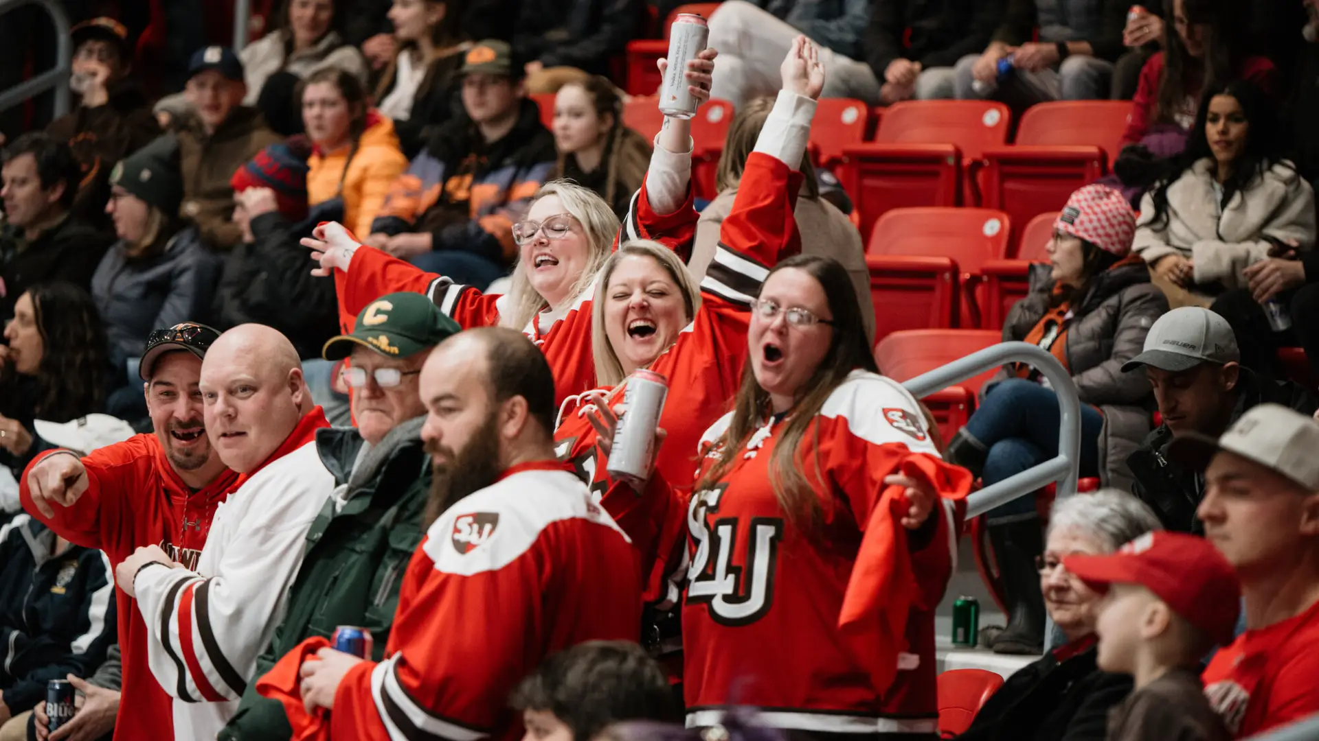 A group of three women in red hockey jerseys cheer and look toward the camera in the crowded stands of the 1980 Herb Brooks Arena during an ECAC Hockey game. 