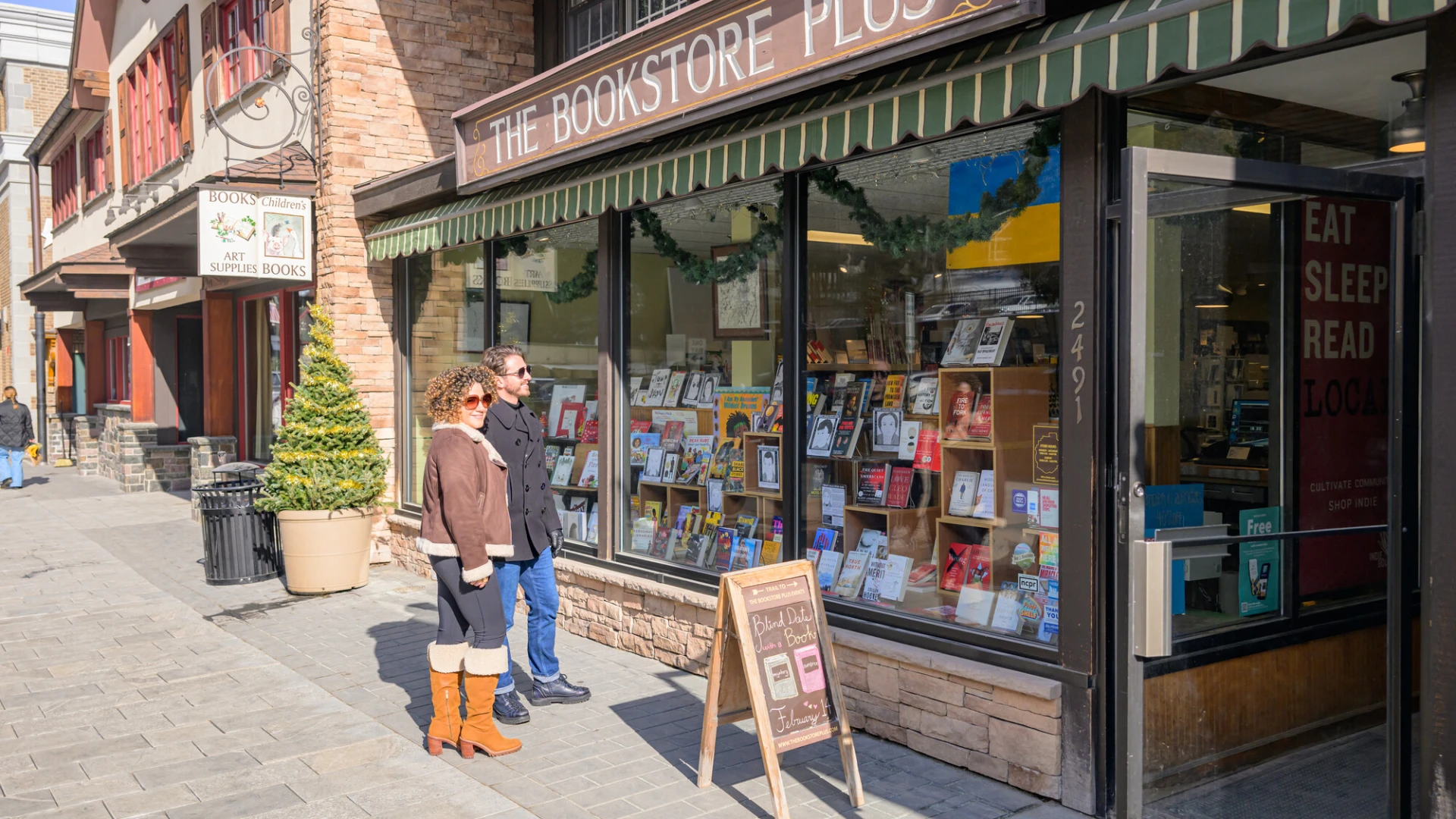 Man in black peacoat and woman in brown leather coat stand outside The Bookstore Plus on Main St Lake Placid