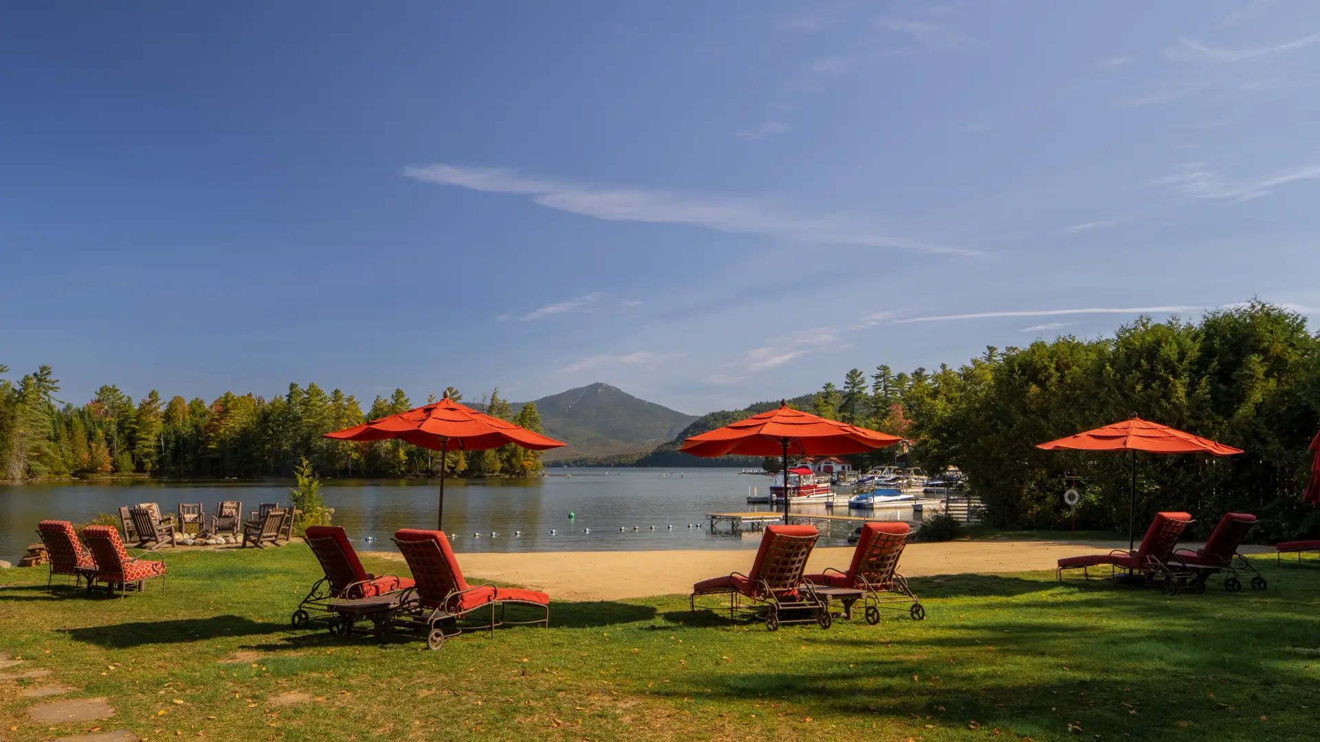 Whiteface Lodge beach on Lake Placid with lawn, sand, beach chairs and umbrellas, boats in the water and a mountain view