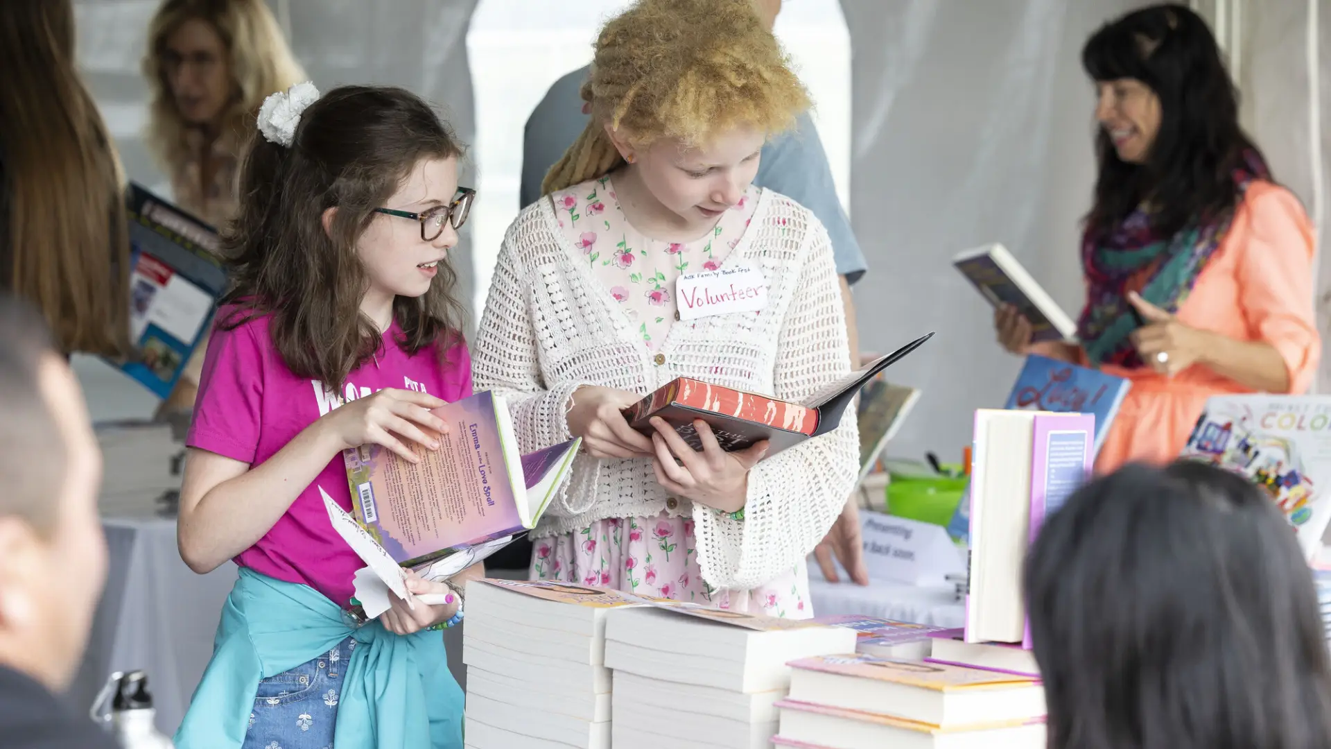 Two girls look at books in front of table stocked with tall piles of books