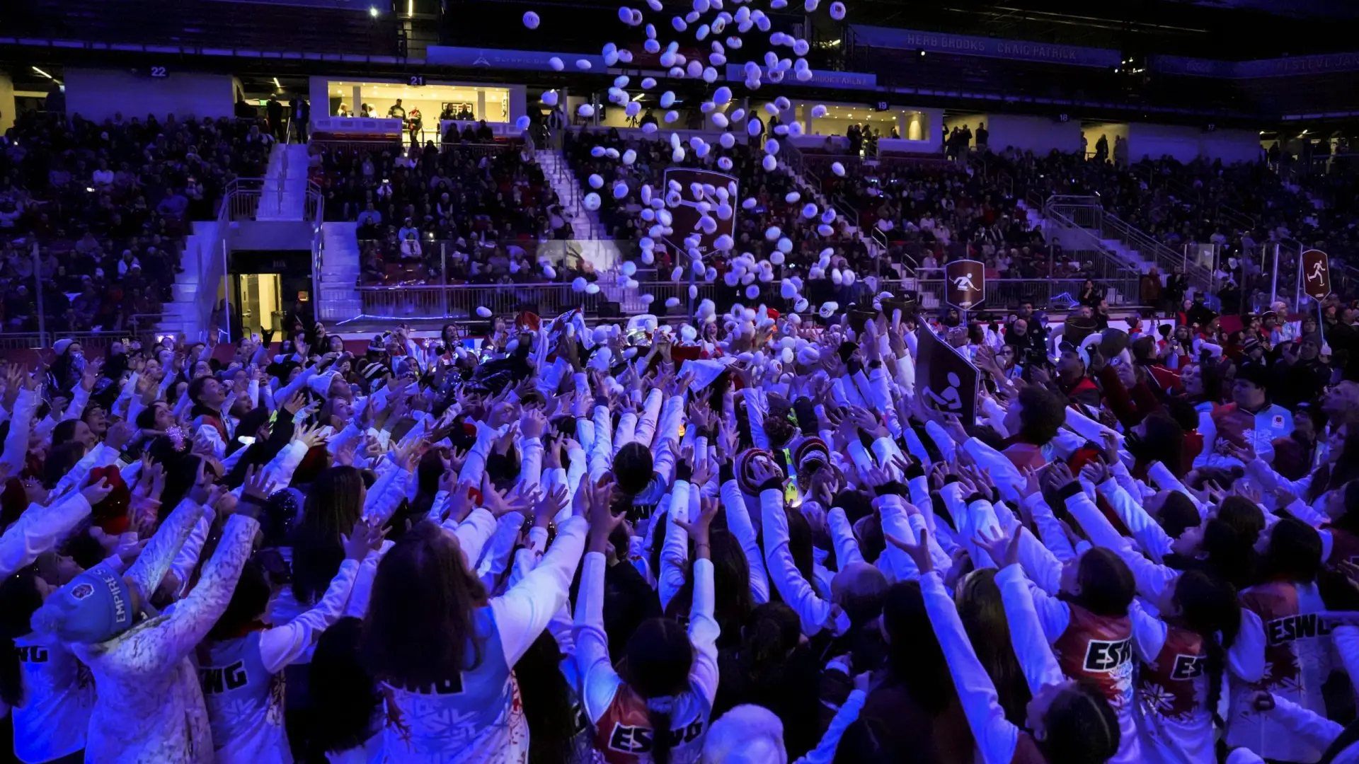 Crowd of people in a dimly lit arena reaching up as white "snowballs" rain down. Excitement fills the scene, with spectators in colorful team uniforms.