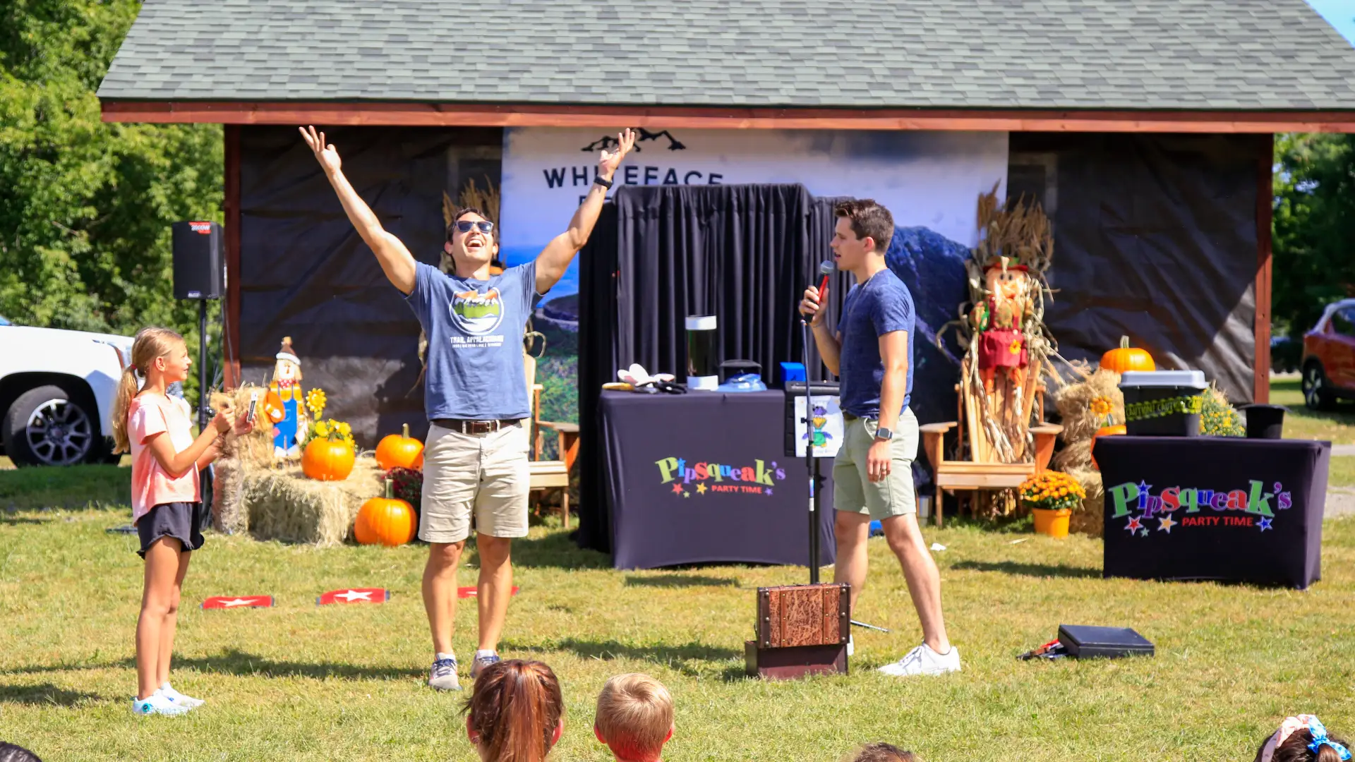 Two men stand at front of crowd of children sitting on grass at fall event, one talking on a microphone and the other raising both arms