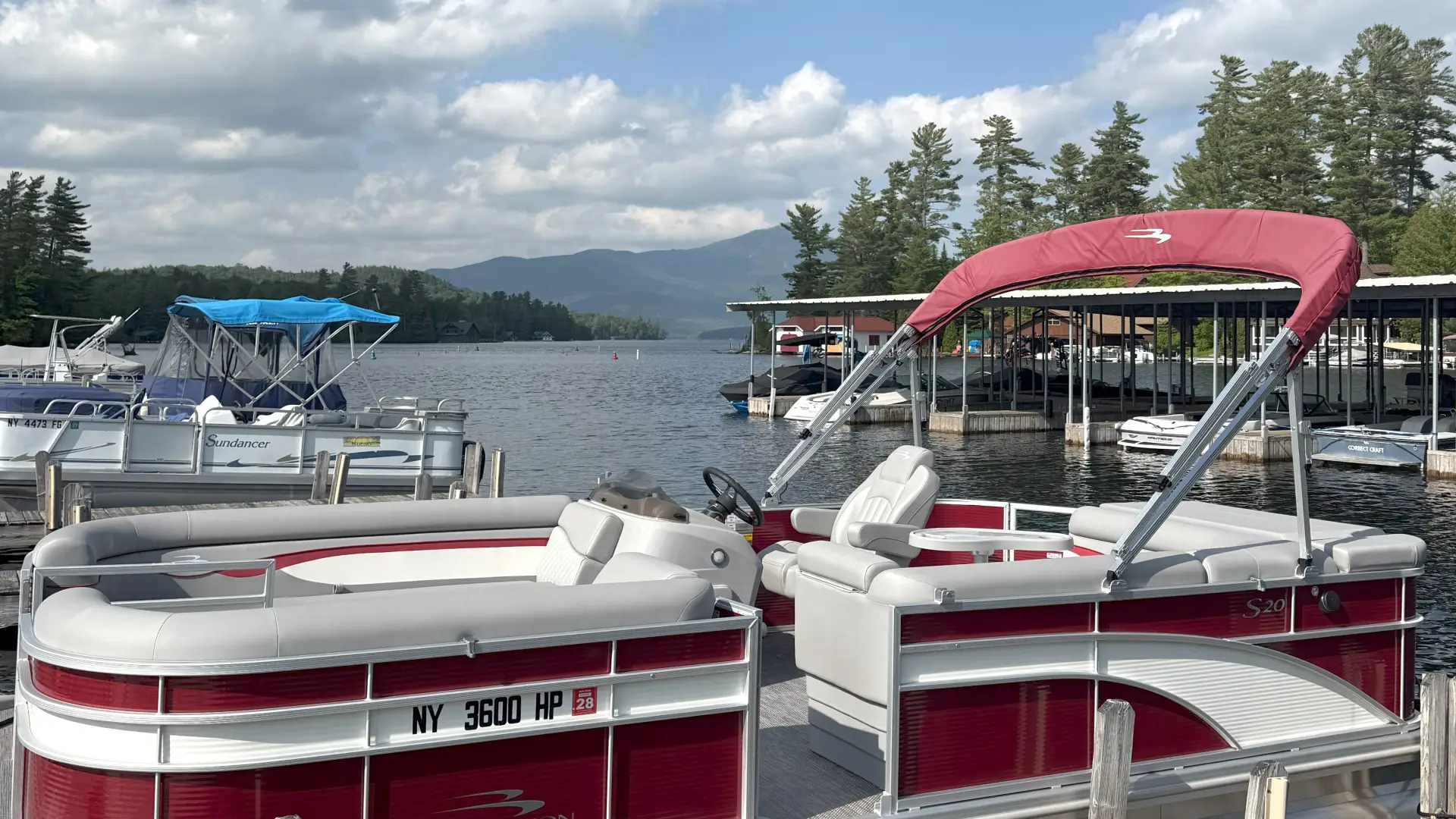 A red and white pontoon boat parked at the marina with a mountain surrounded with white billowing clouds