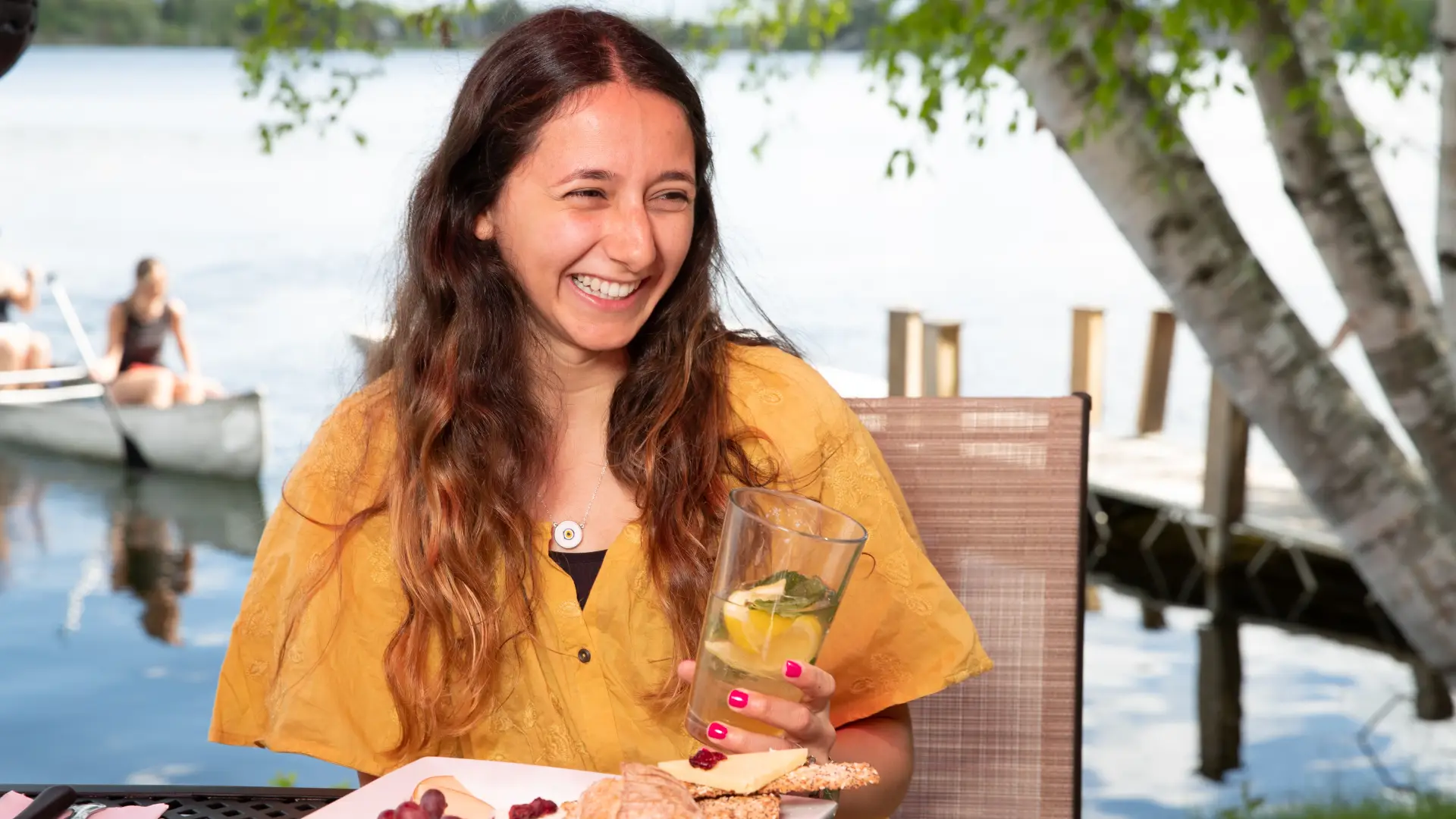 A young woman smiles widely at a table of food, while holding a drink in front of a lake with canoeists.