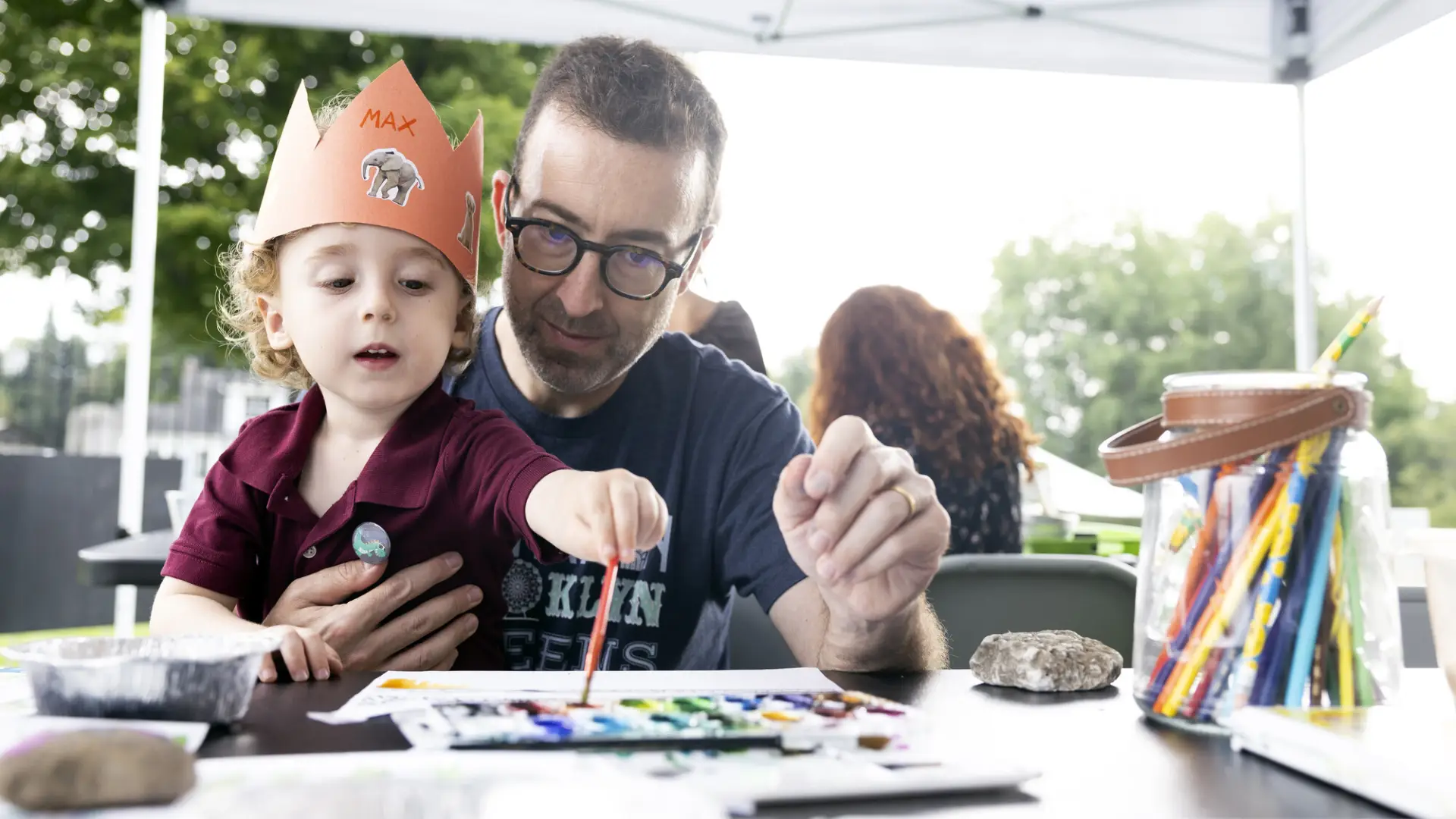 Little boy wearing paper crown sits on man's lap dipping paintbrush in watercolors