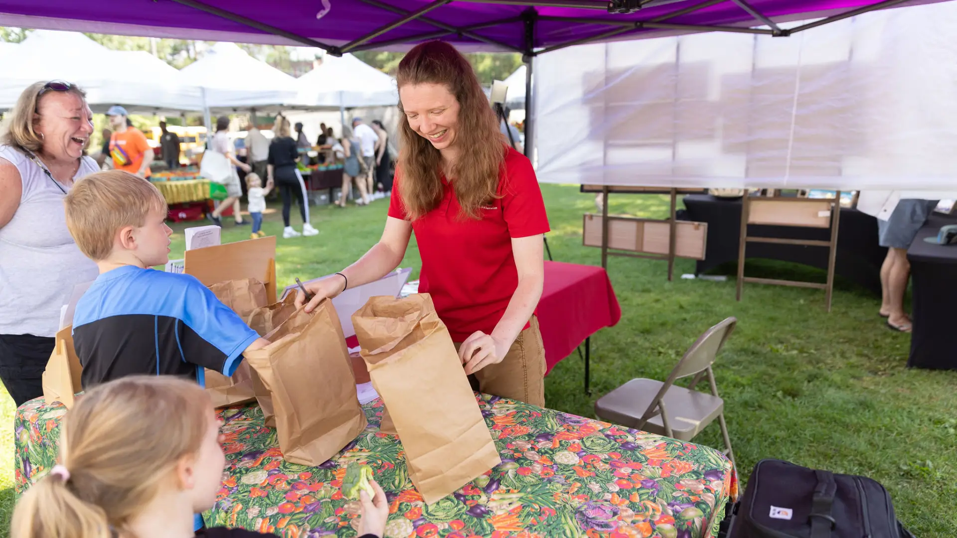 A woman at a farmer's market fills brown paper bags with produce for children's activities.