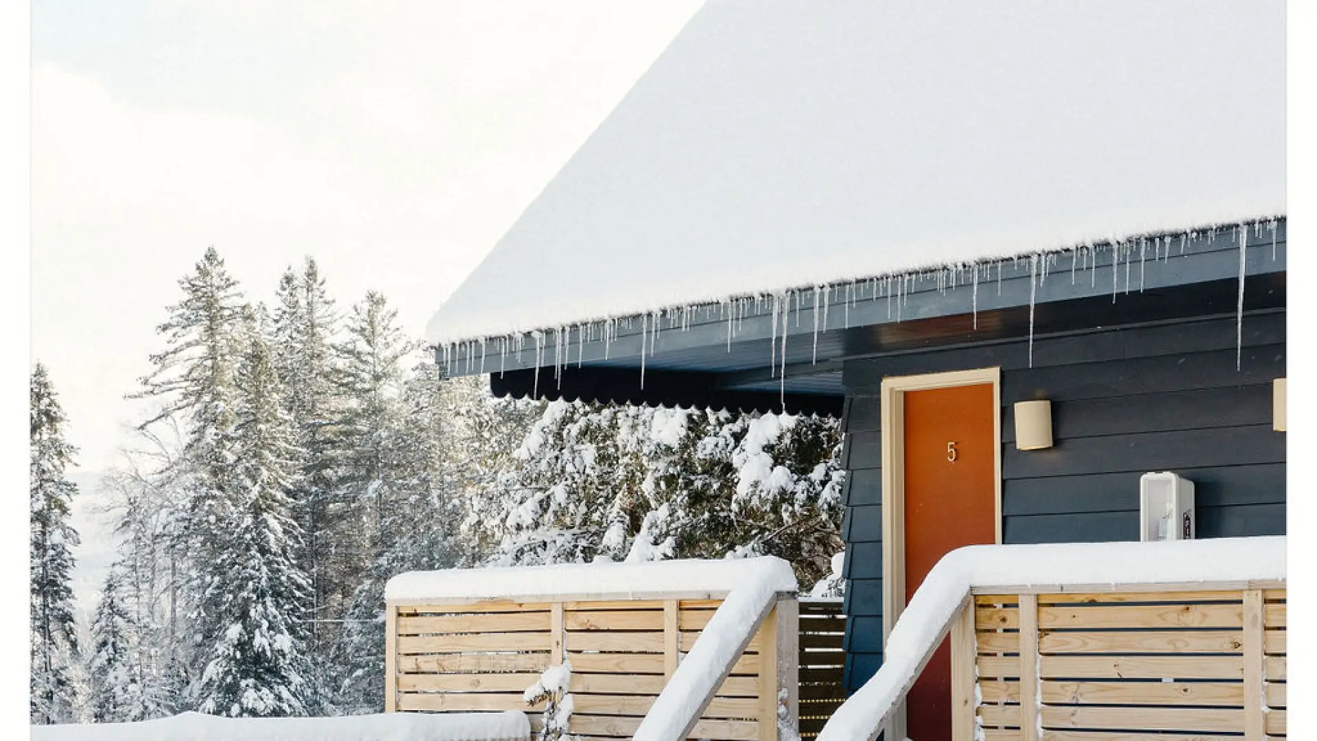 The exterior of one of the cabins with large porch blanketed in snow with tall pines in the background.