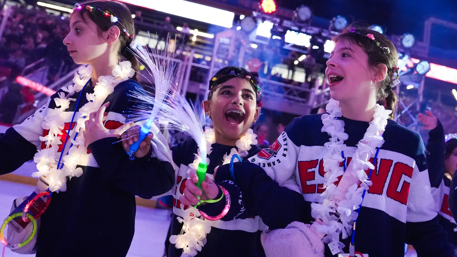 Three smiling children wearing "ESWG" sweaters celebrate with light-up wands during an opening ceremony.