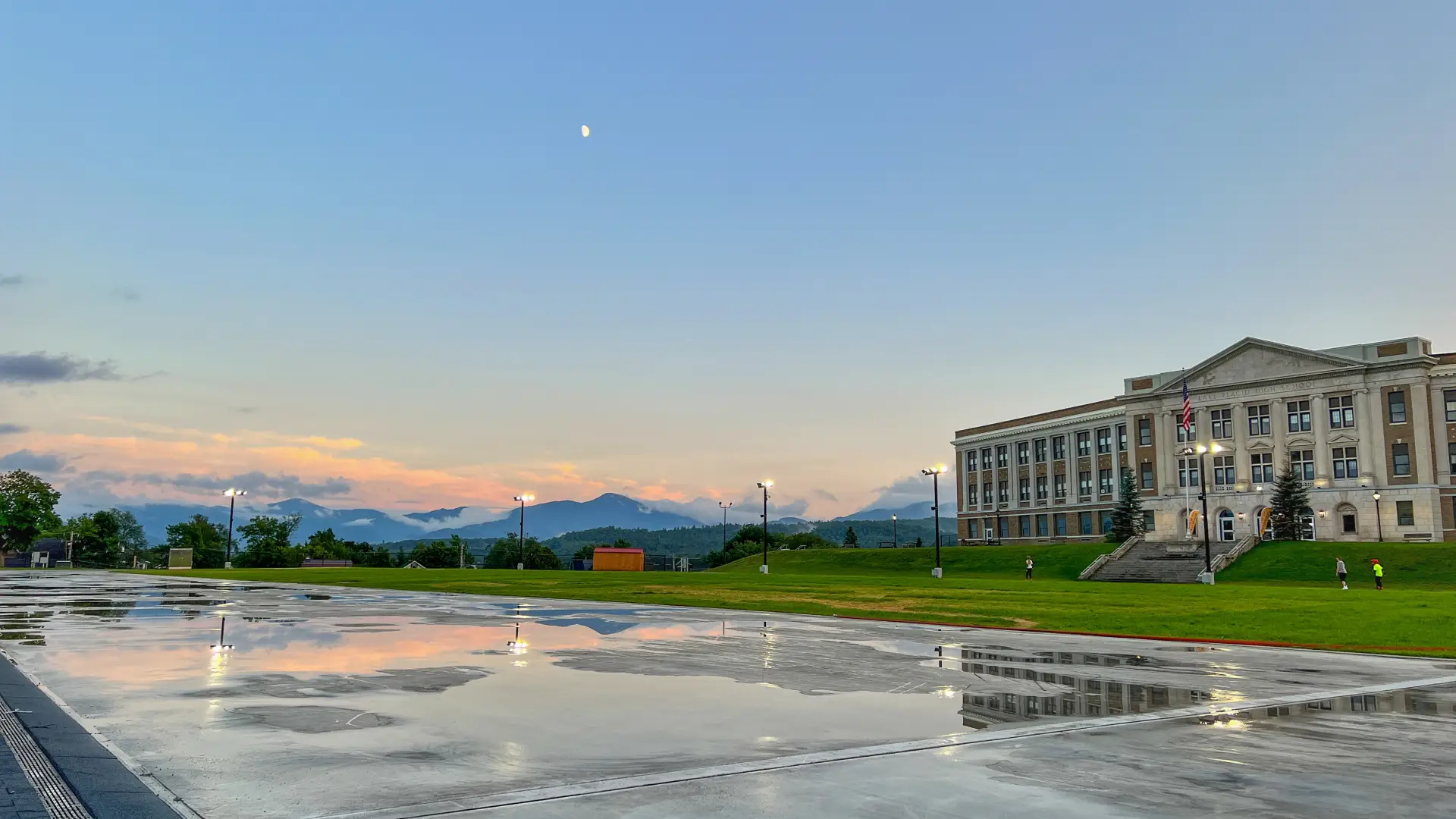 Puddles of rain glisten on a paved outdoor skating track. The moon gleams in the dim blue sky above.