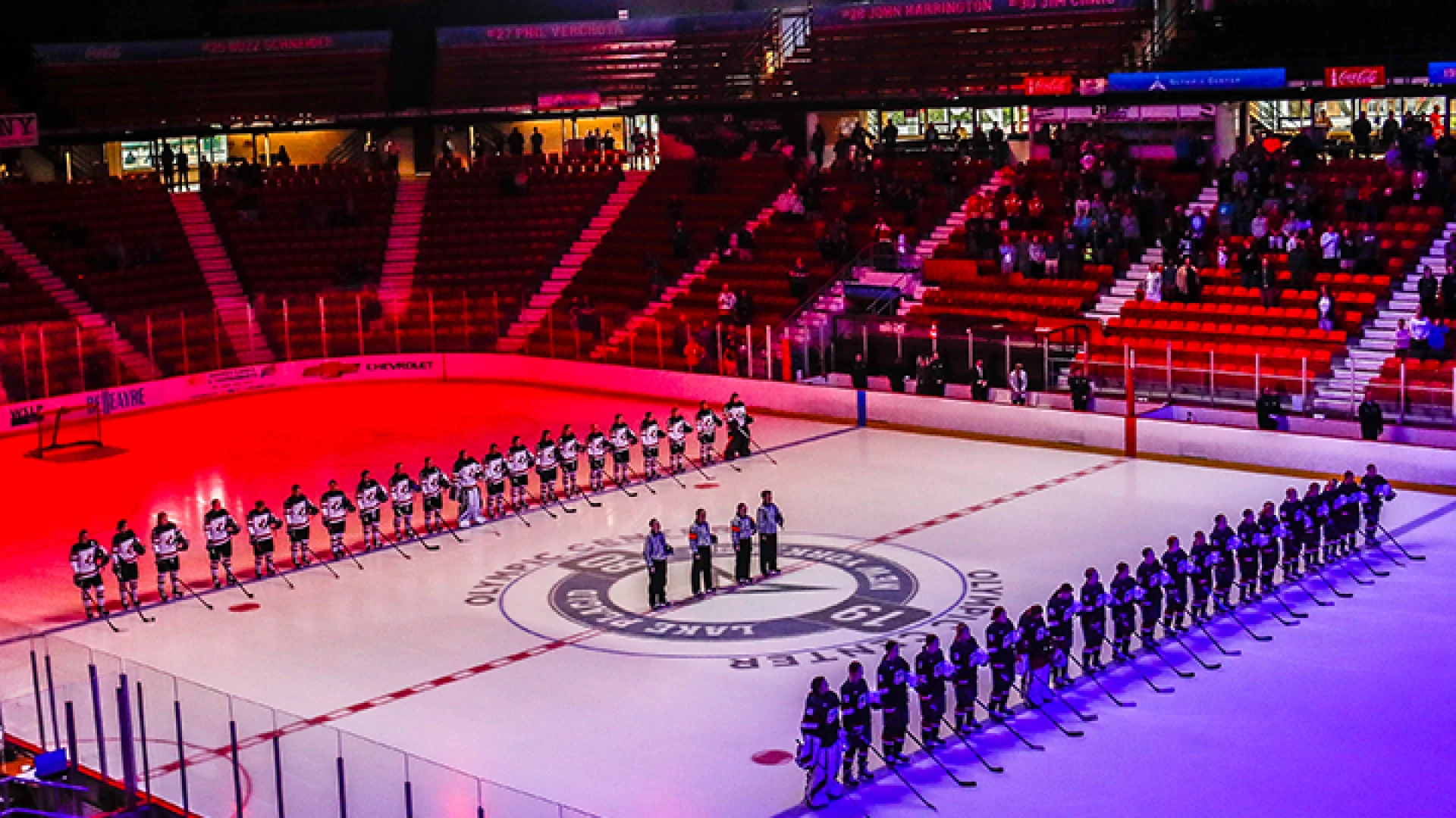 Two teams of female hockey players lined up opposite one another on the ice with officials in the middle under colorful light