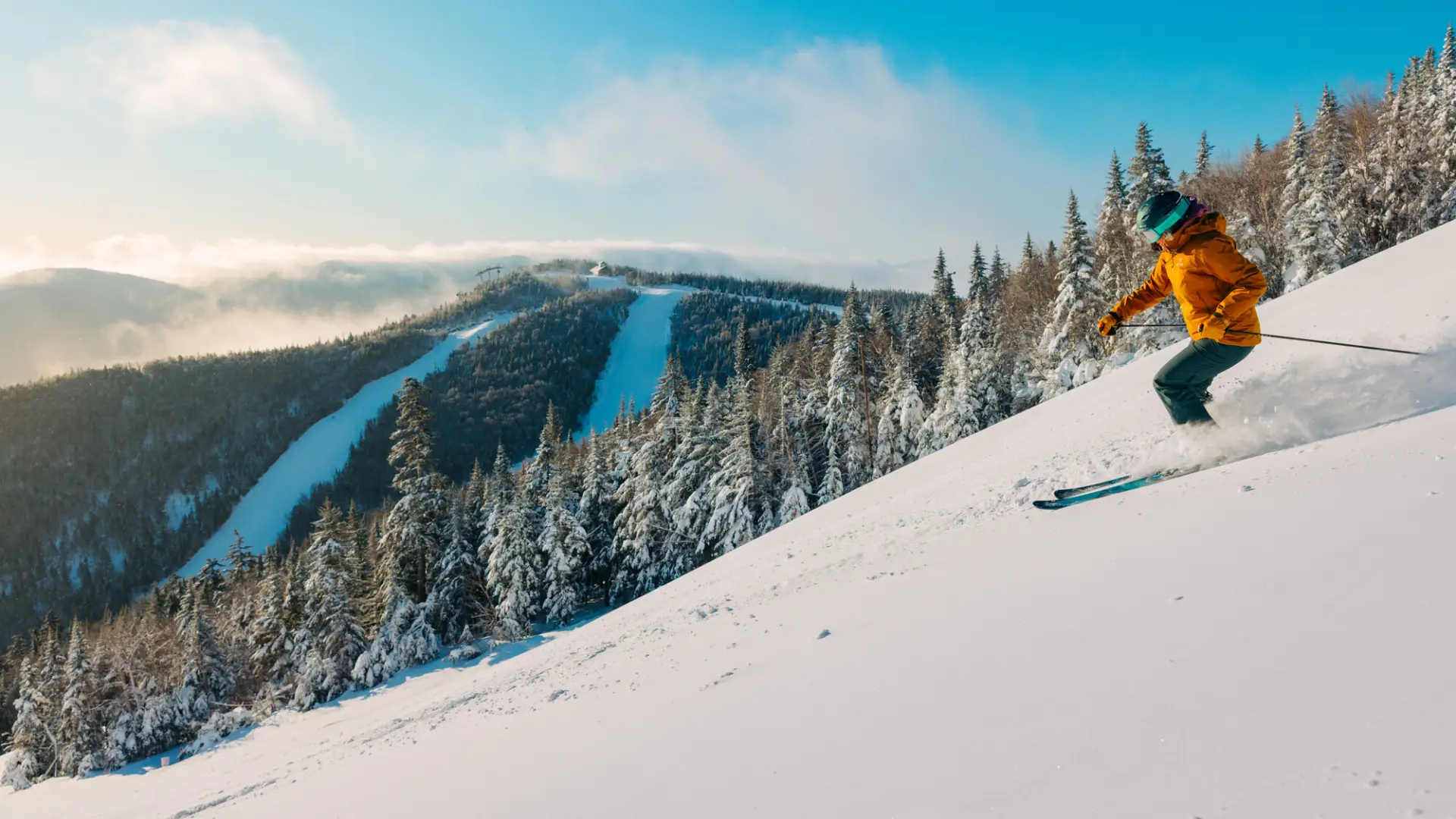 Person in orange ski coat and ski gear descends snowy slope on largely blue sky day with additional mountain peak with snowy runs in bakcground