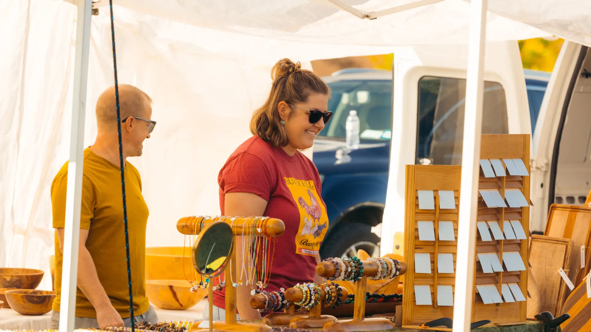 Man and woman browse table of jewelry under white tent at farmers market