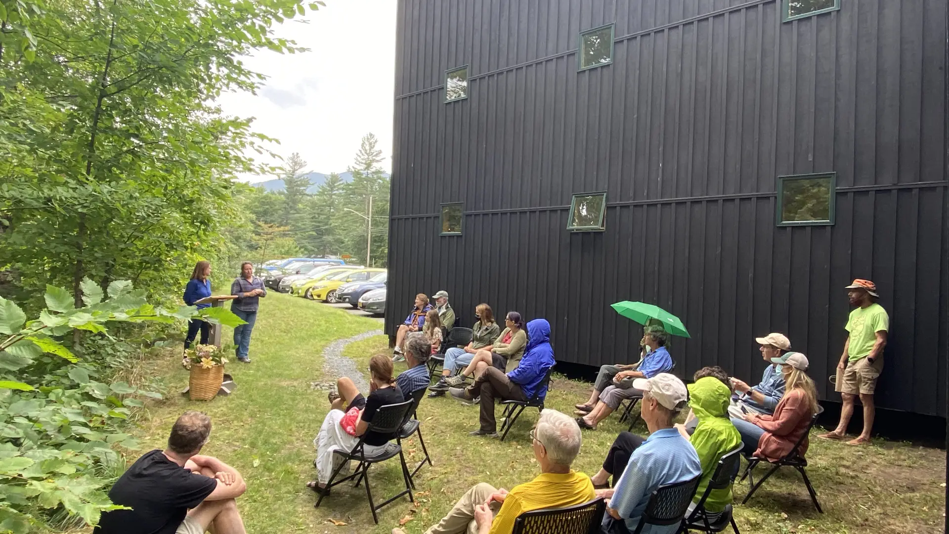 A group of people are seated in chairs outdoors, listening to two people giving a presentation.  A dark building is in the background.