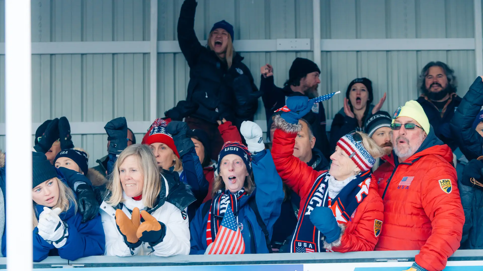 Crowd of people in winter attire stands under awning next to luge track cheering