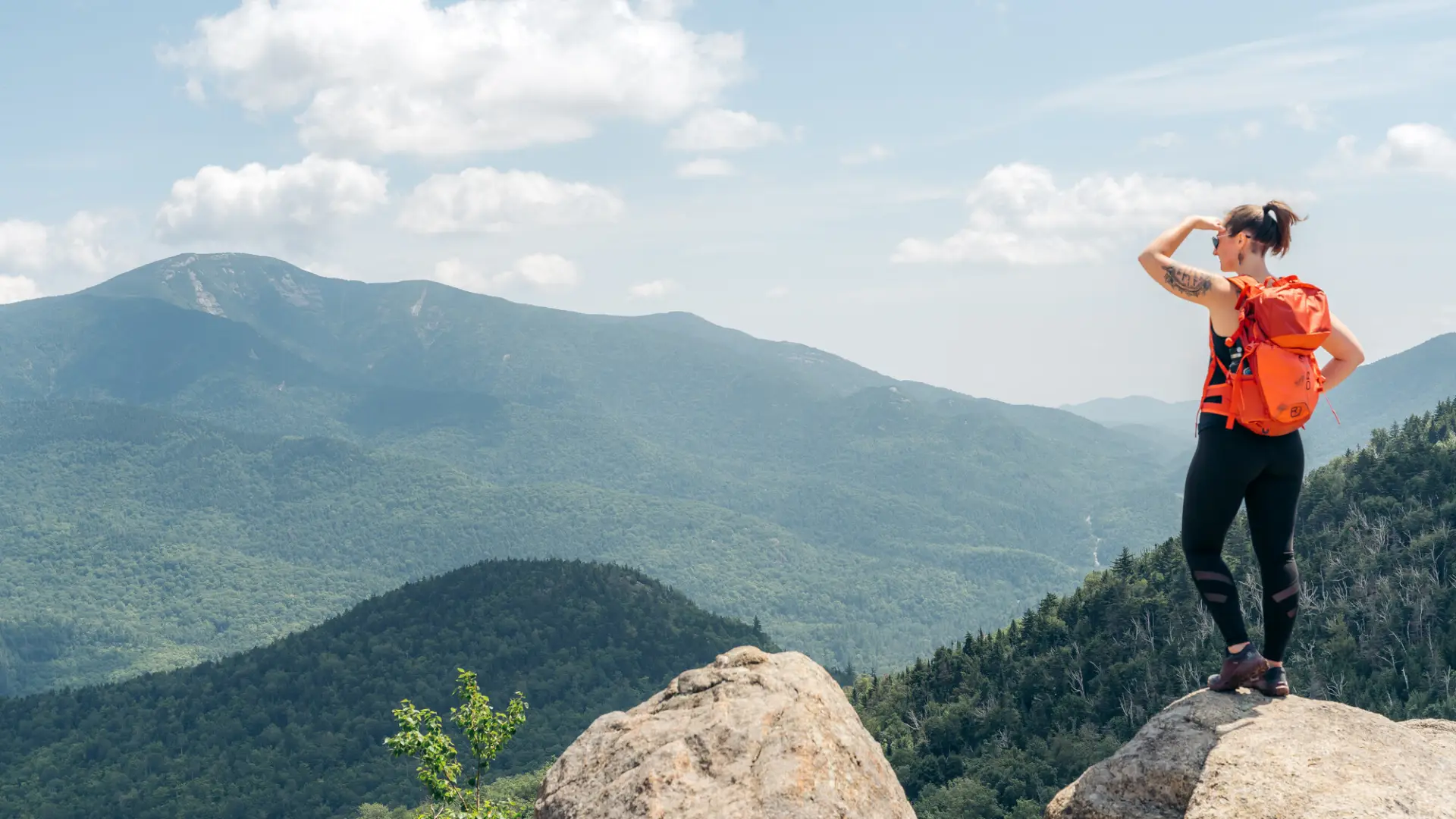 A woman standing at the summit of a hike, looking out at the mountain views.