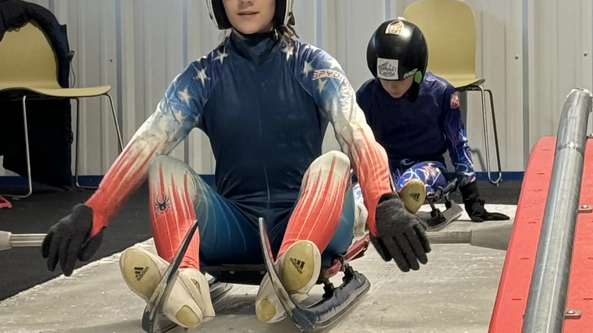 A young woman in a USA luge uniform sits on a luge sled on an indoor track.  Another young person is visible behind her, also on a sled.