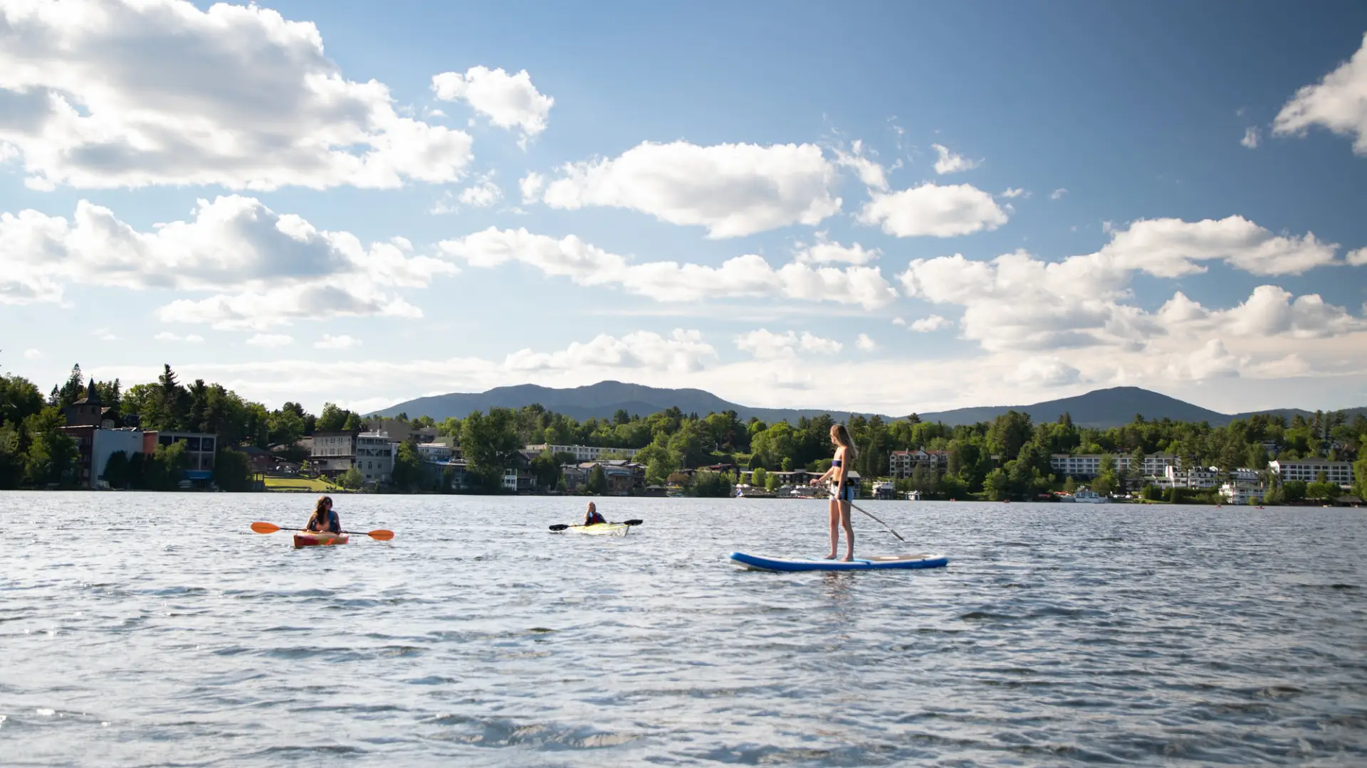 Kayakers and a woman on a SUP relax on a lake with town buildings in the background.