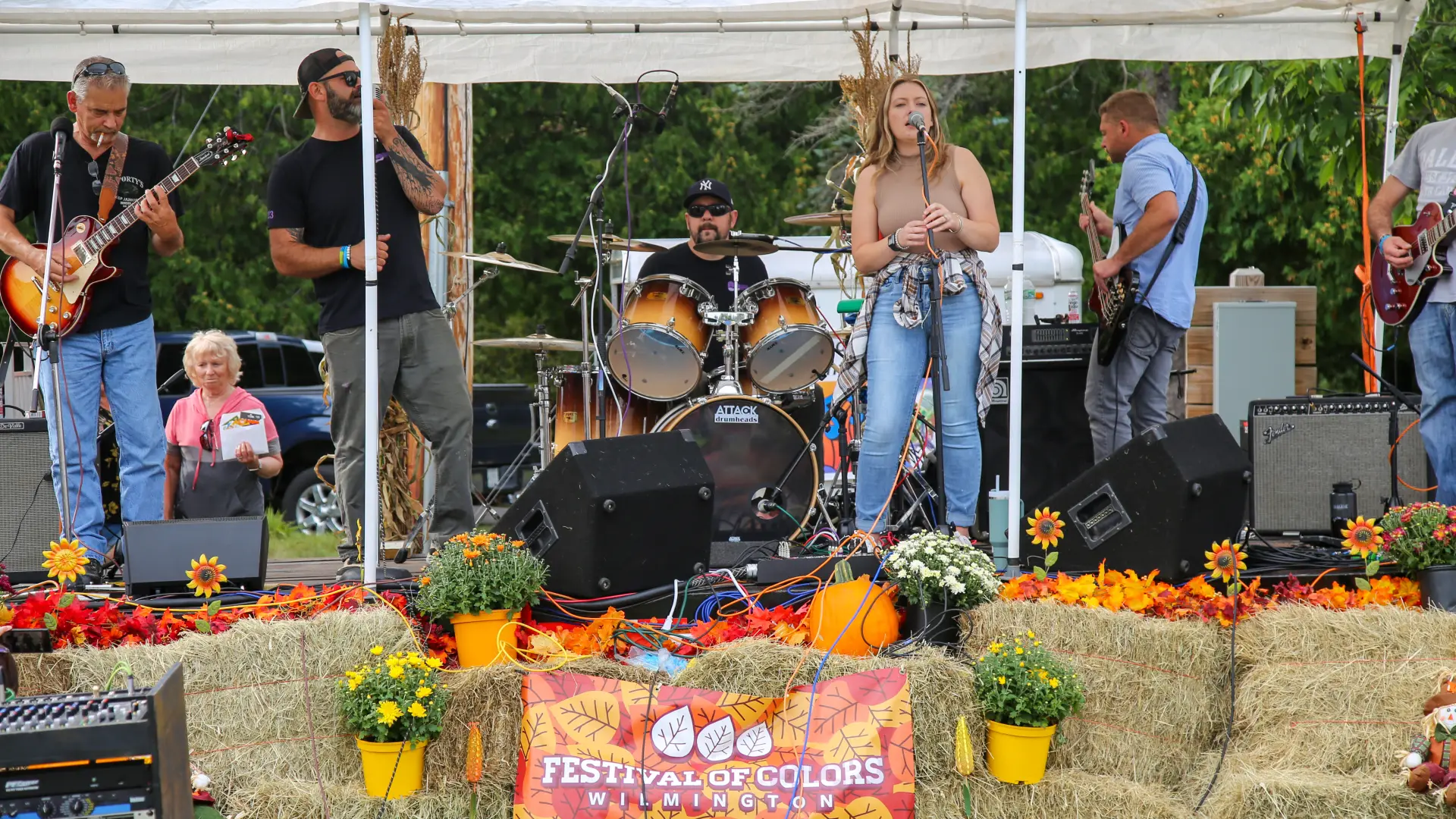 Band with female lead singer performs on outdoor stage under white tent with hay bales, pumpkins, and mums decorating a stage with a small Festival of Colors banner