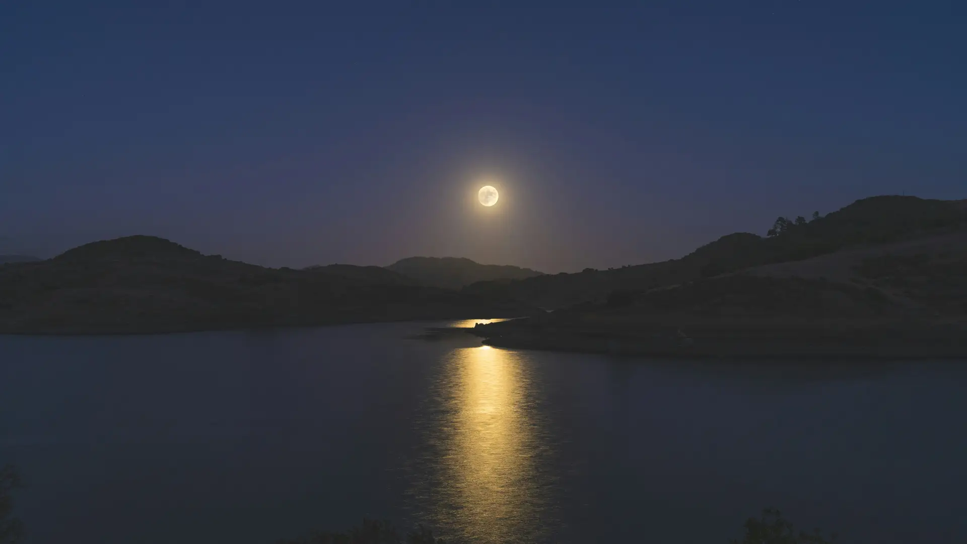 Full moon in dark sky over lake and trees shoreline hills with moon reflecting in the water