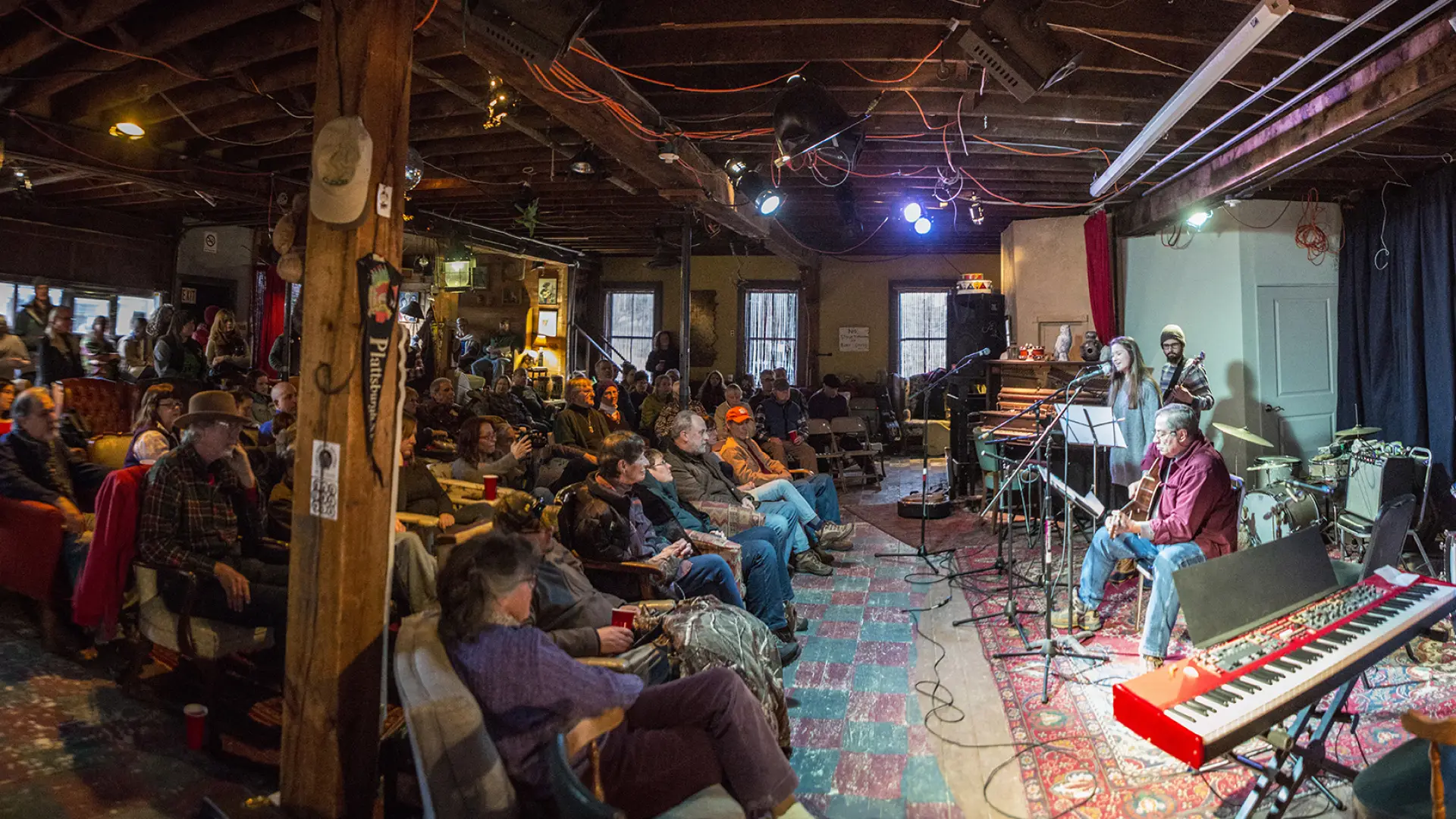 Man plays guitar on stage at Upper Jay Art Center with full house of people sitting on various chairs and couches