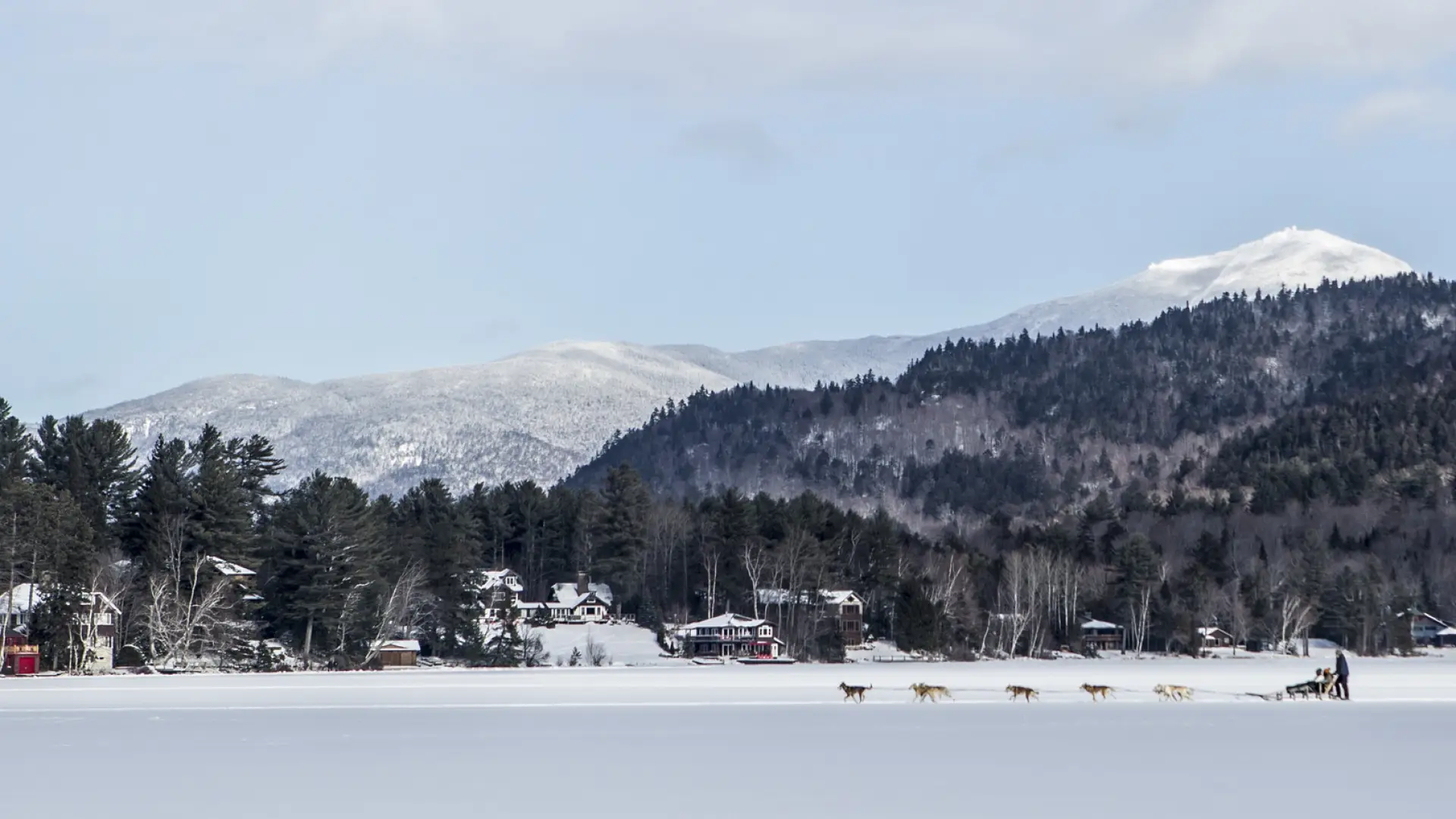A dog sled team races across a frozen lake with mountains in the background.