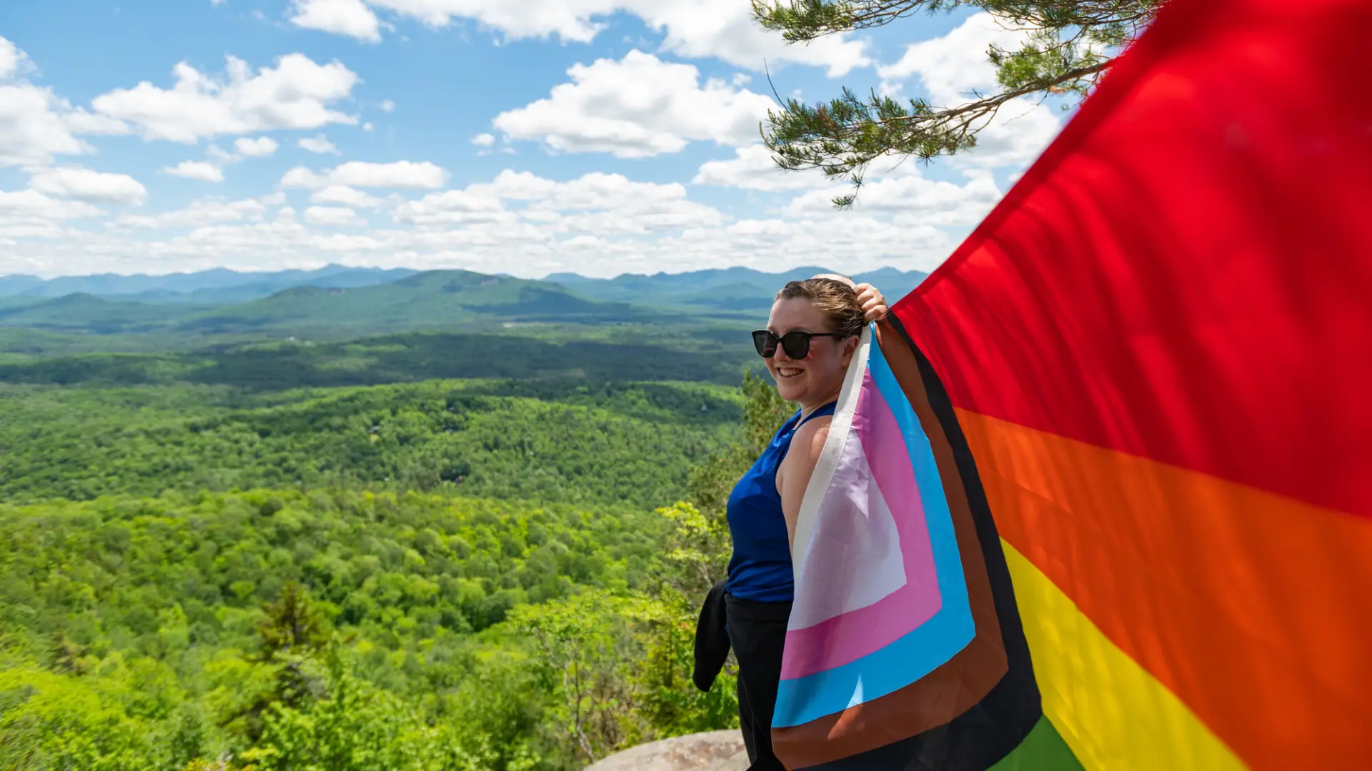 Person in hiking clothes and sunglasses stands on top of a mountain in summer holding pride flag billowing behind them