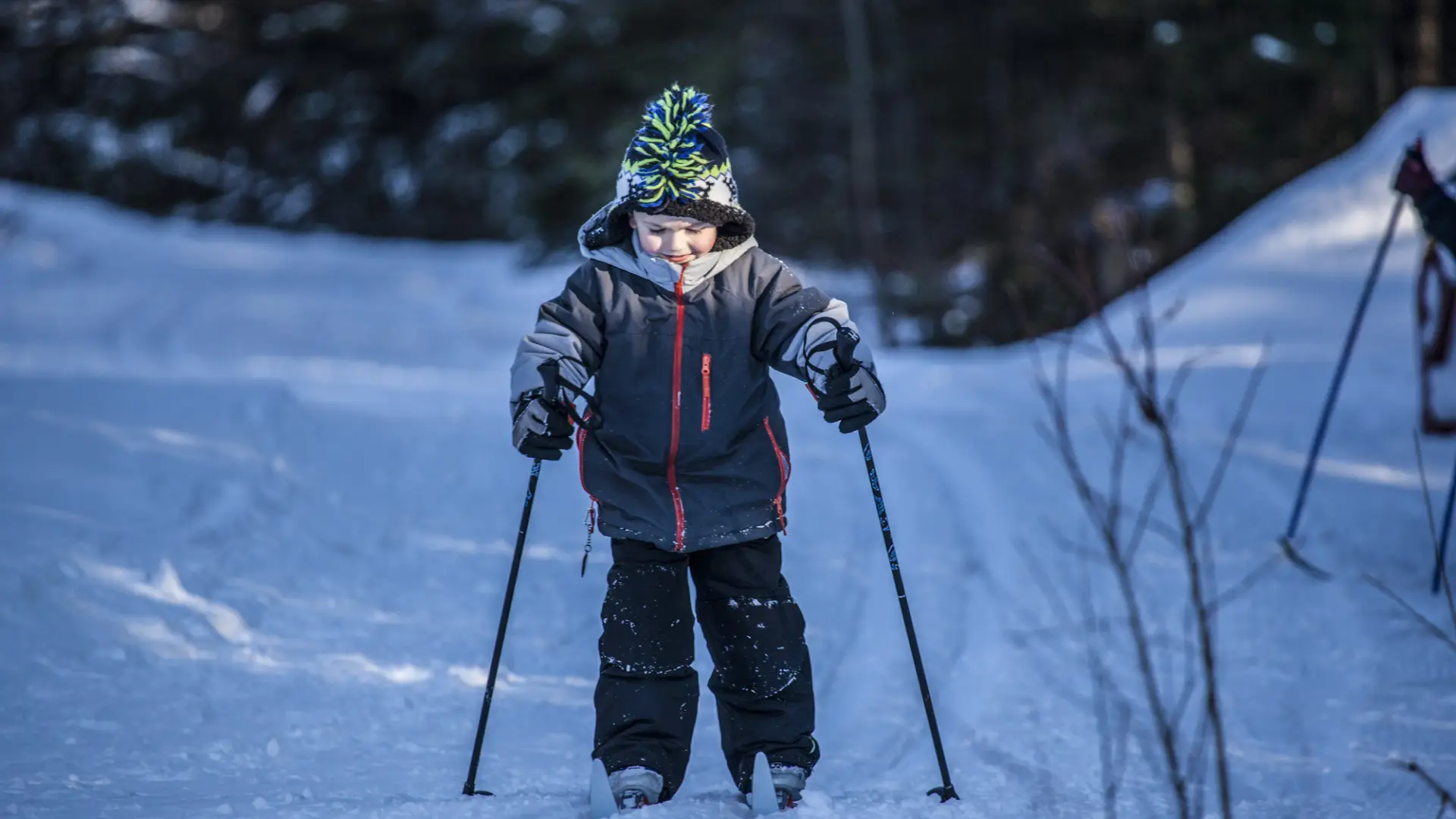 A young boy smiles and cross-country skis on a groomed trail in the woods