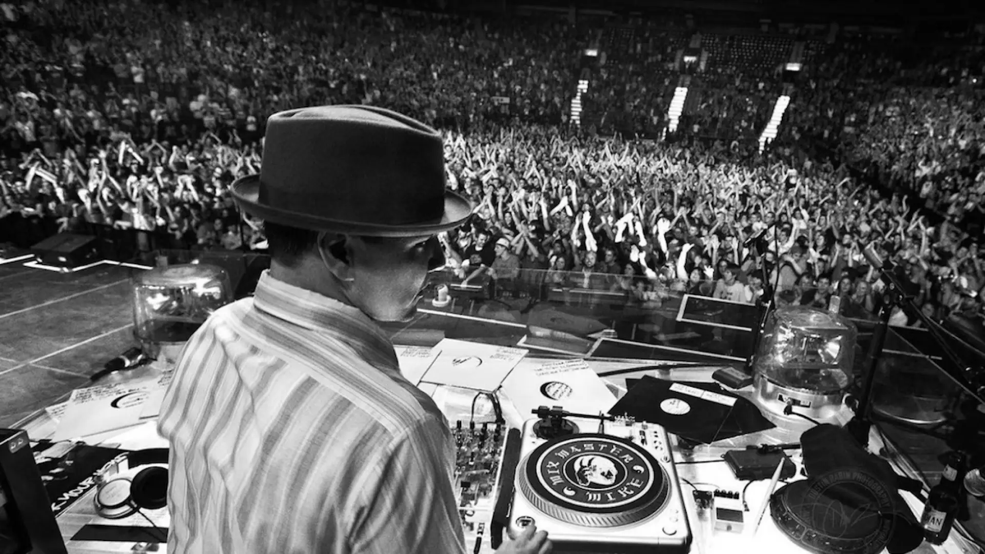 A black and white photo of a DJ wearing a fedora in front of turntables at a concert.