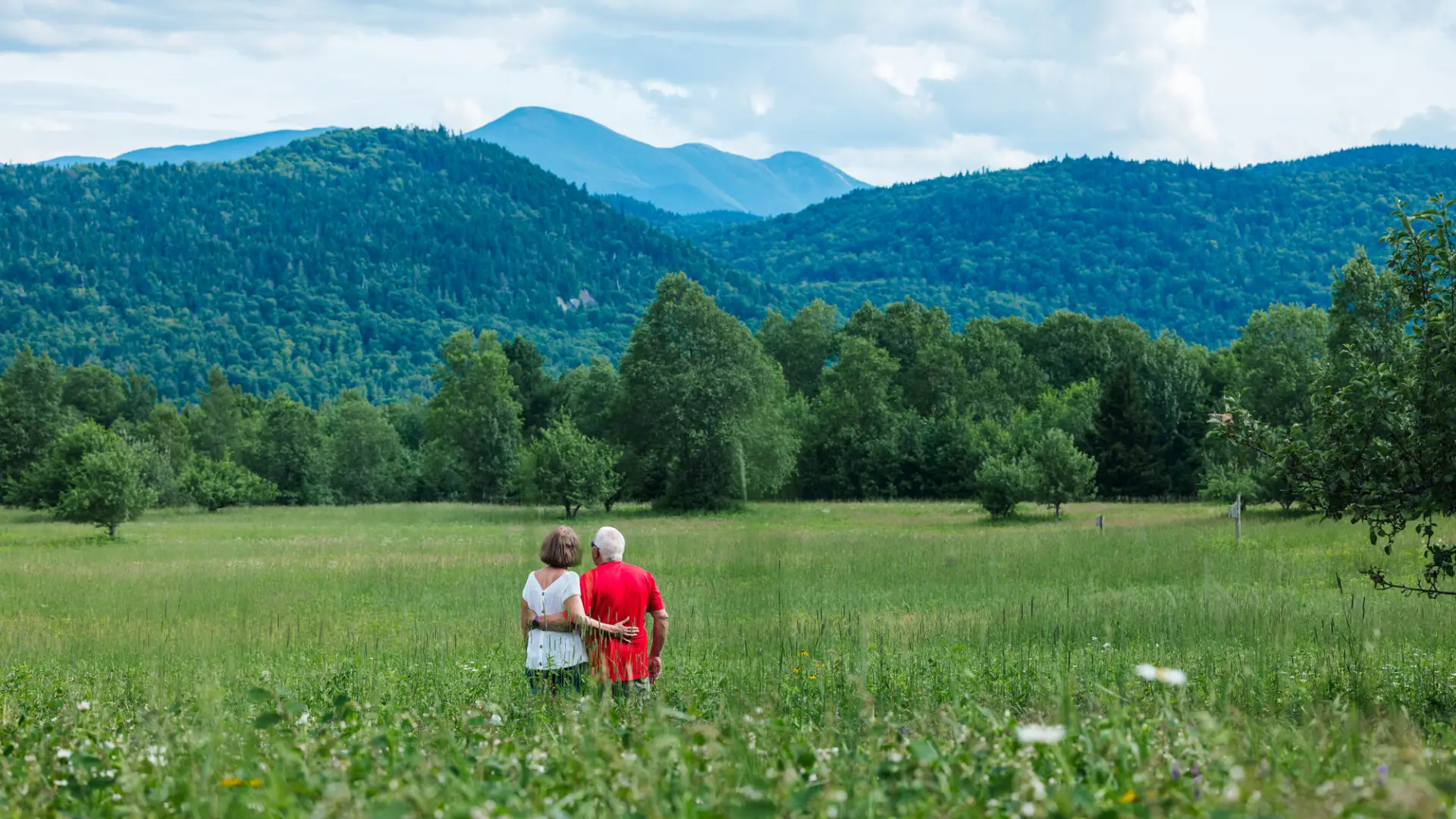 Senior couple stands in field of tall grass with arms around each other's waists looking at mountain view