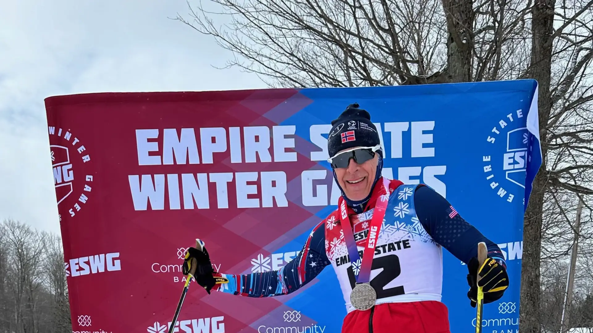 Skier with medals and ski poles poses proudly in front of a branded Empire State Winter Games backdrop, wearing a red vest and blue tights on snow.