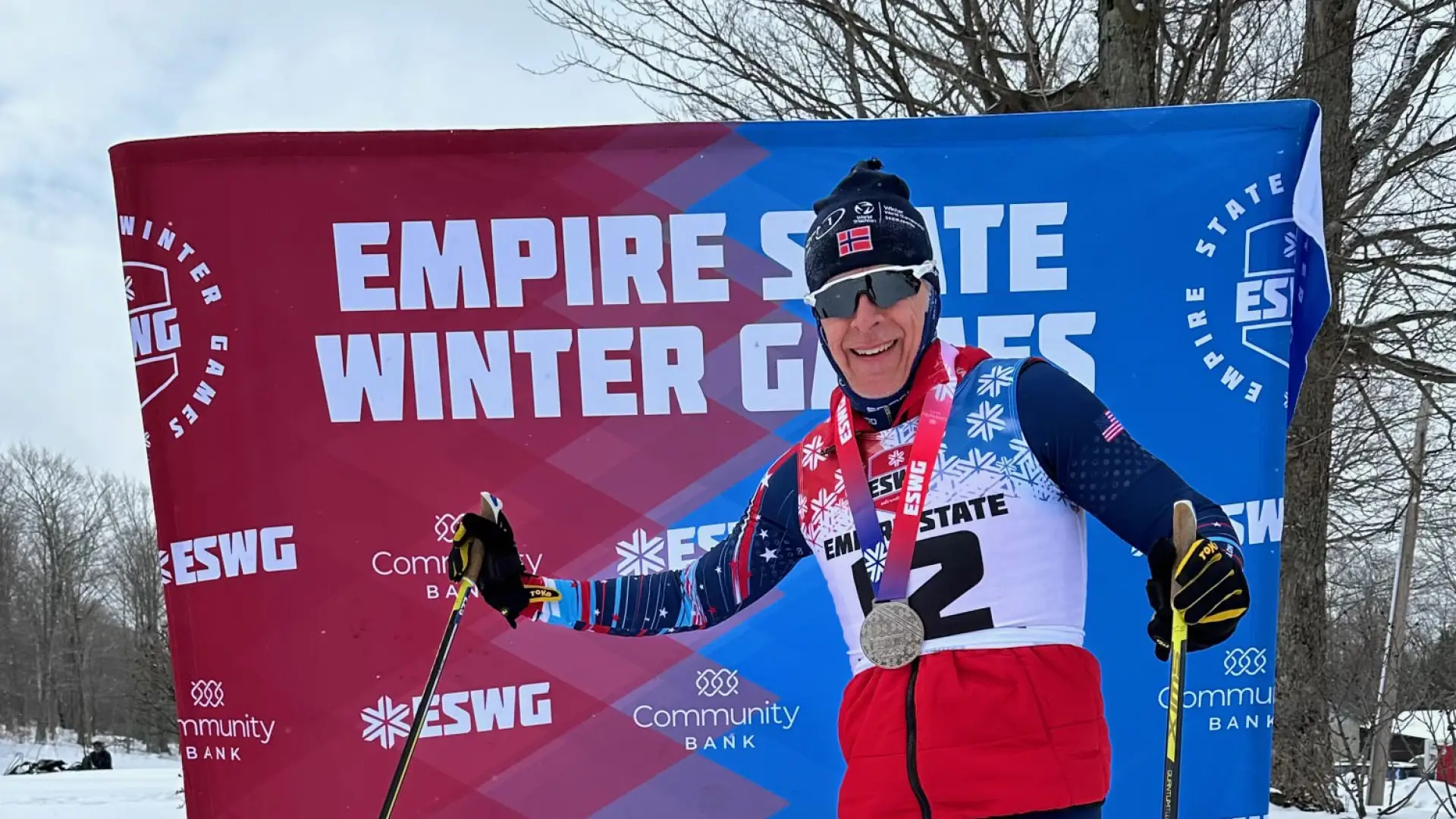 Skier with medals and ski poles poses proudly in front of a branded Empire State Winter Games backdrop, wearing a red vest and blue tights on snow.