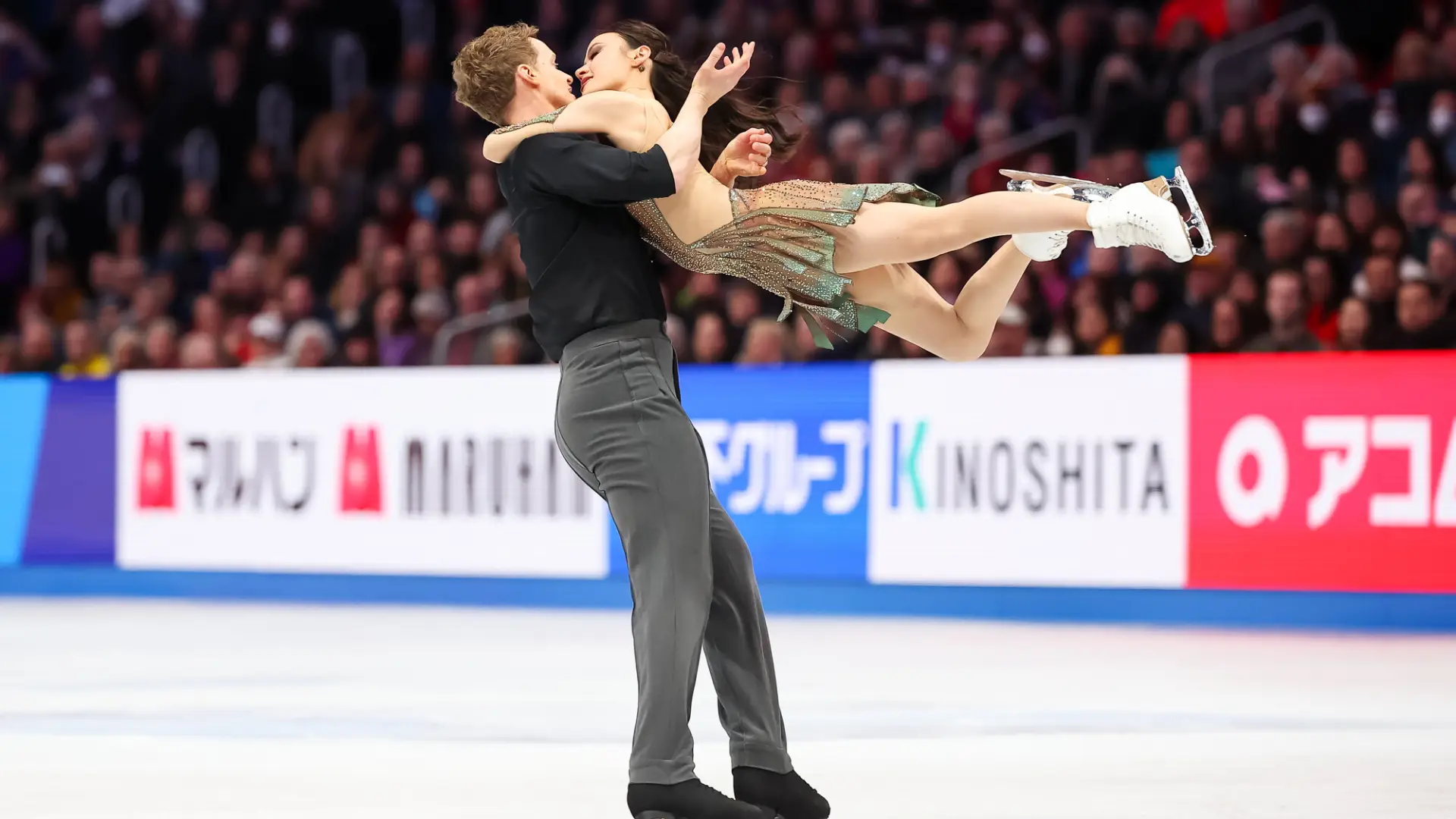 Male figure skater holds up female figure skater while performing on ice with a large crowd watching