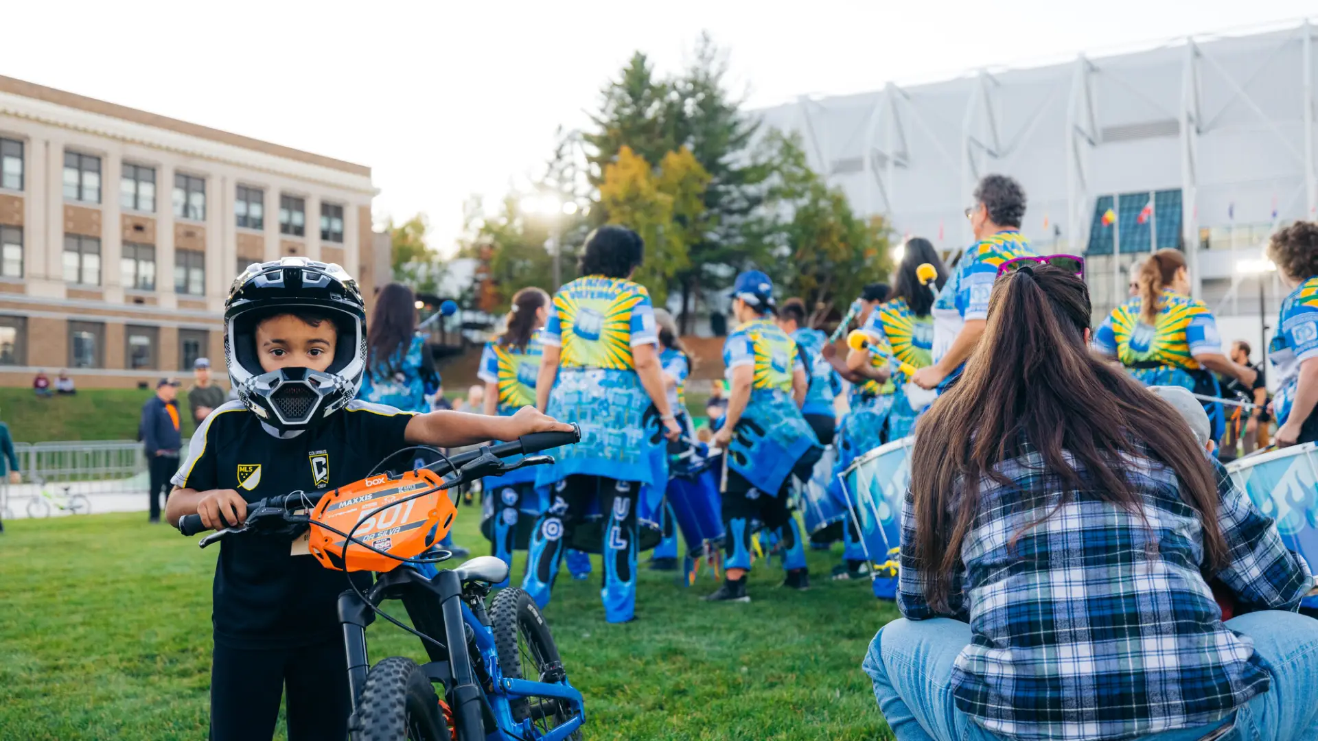 Young child looking at camera holds small bike and wears full Mountain Bike helmet in front of drum line performers