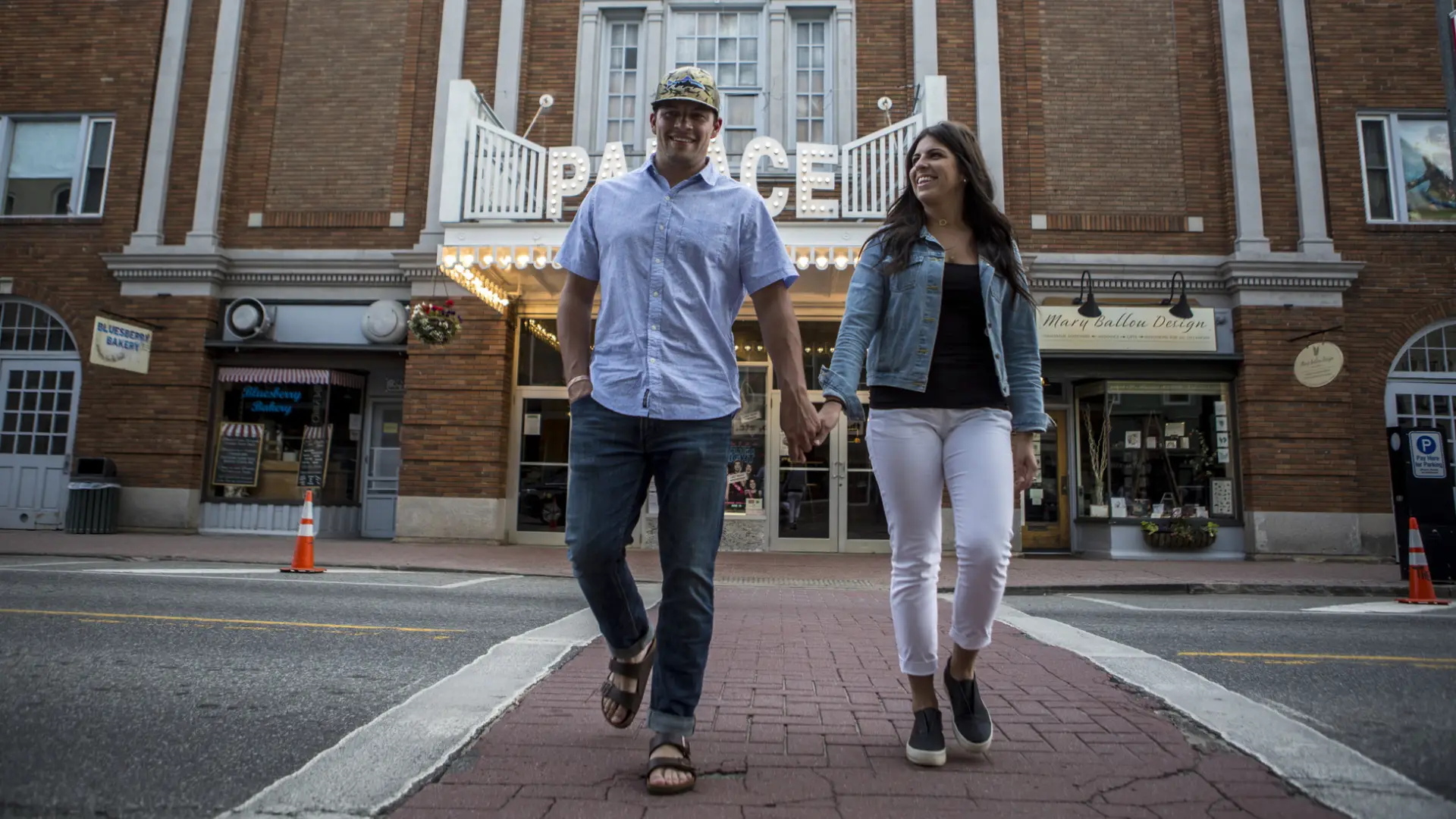 A man and woman hold hands crossing a street with a brightly-lit old fashioned movie marquee behind them.