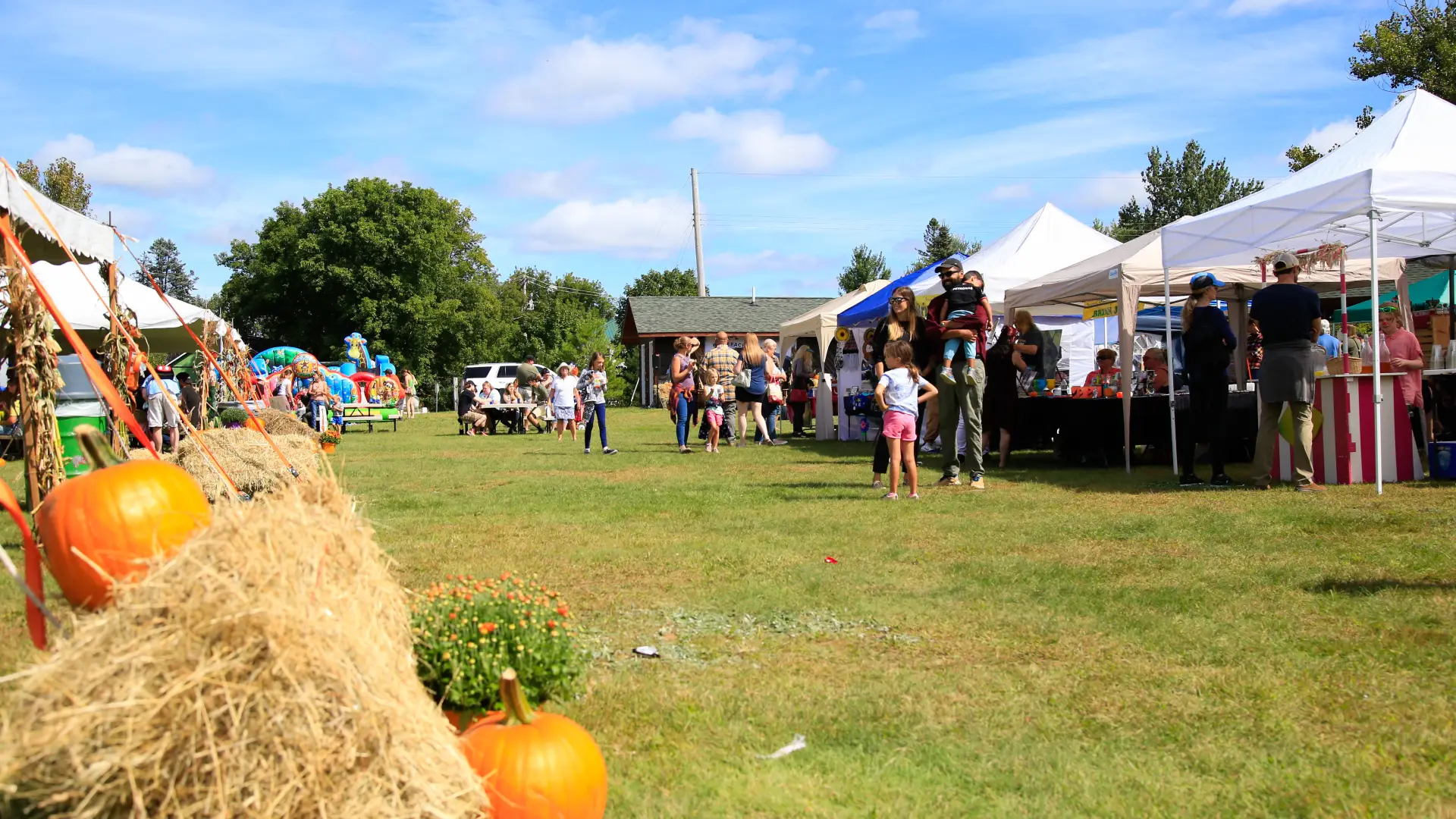 People browse at vendor tents in the background at outdoor event with pumpkin and hay bales in foreground