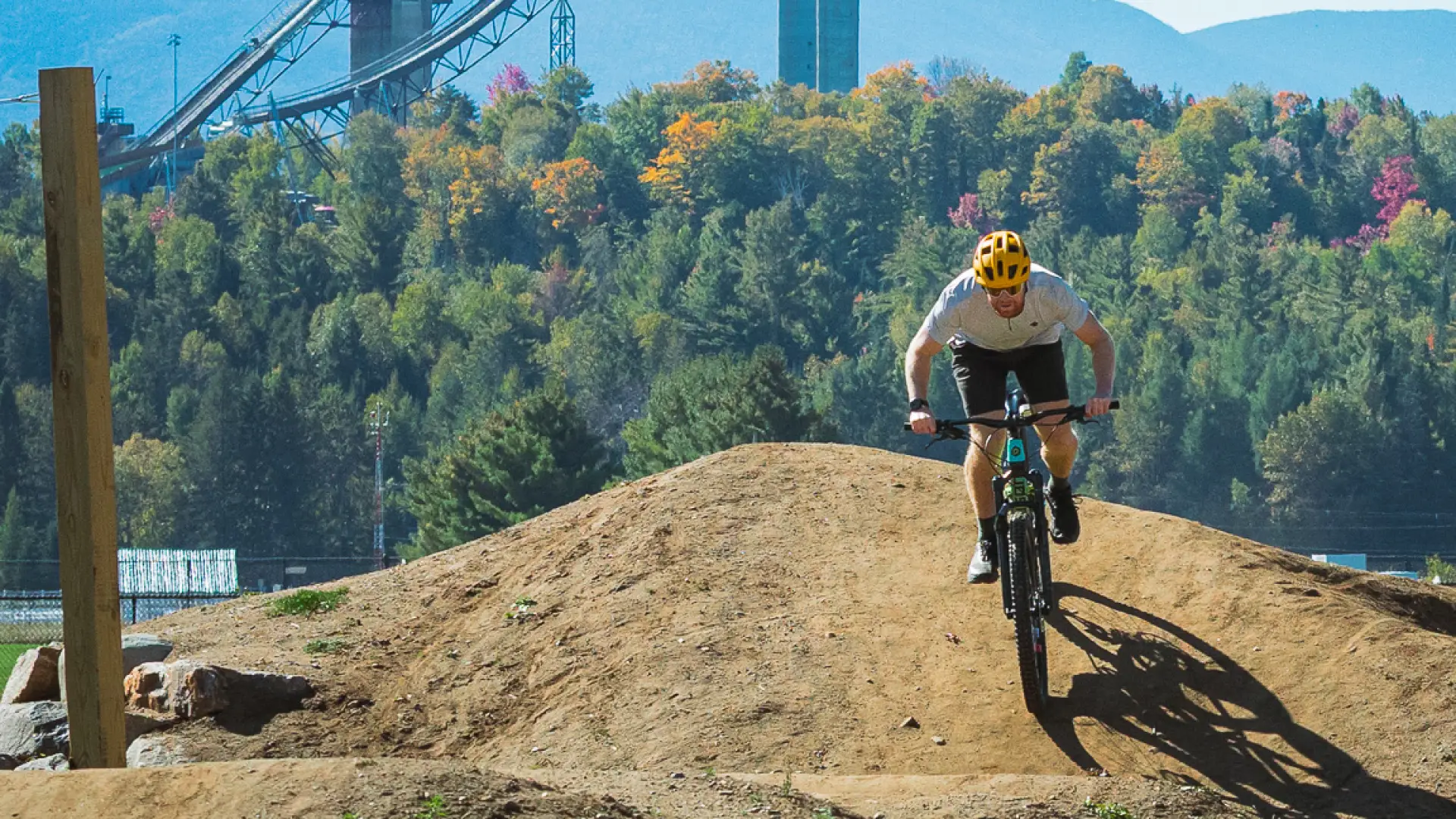 A man rides a mountain bike over a dirt hill with mountains in the background.