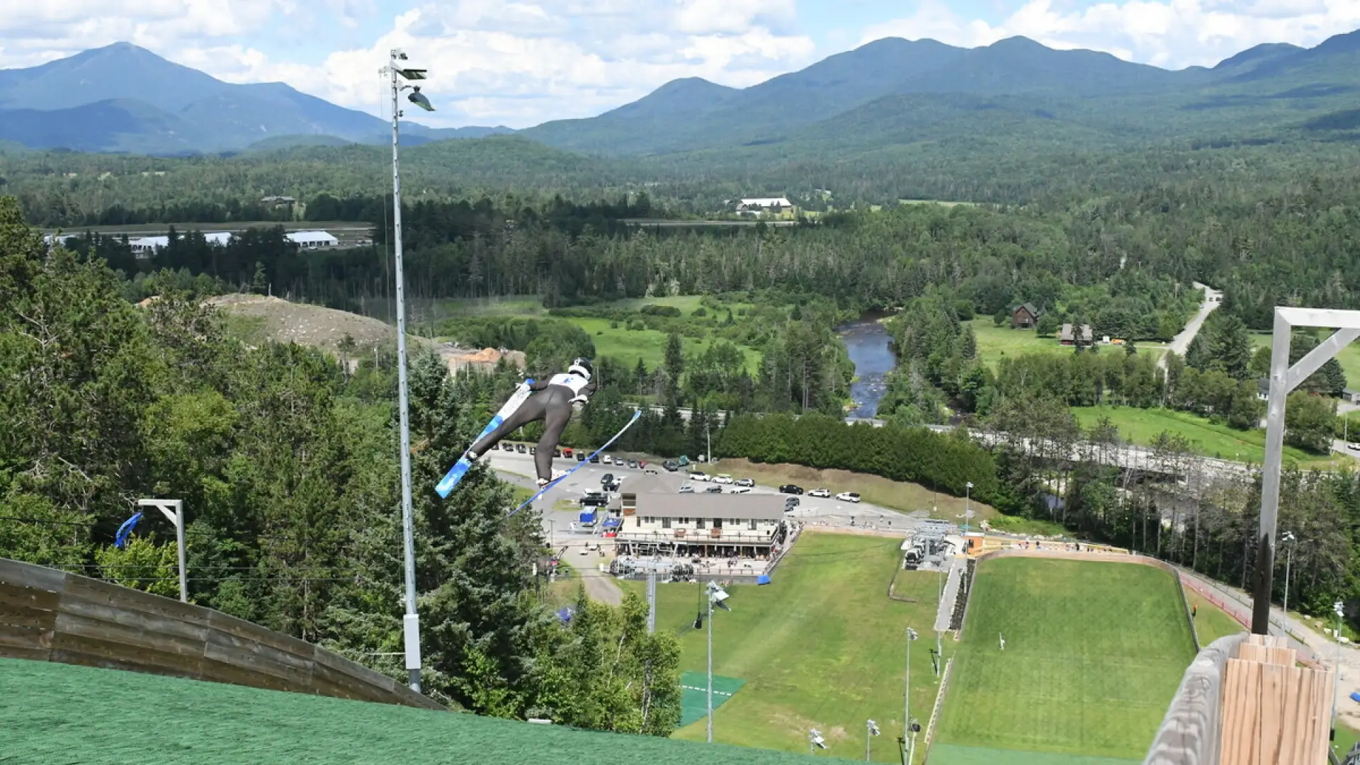 Ski jumper seen flying through the area above turf jump track in summer with mountains and venue in the distance