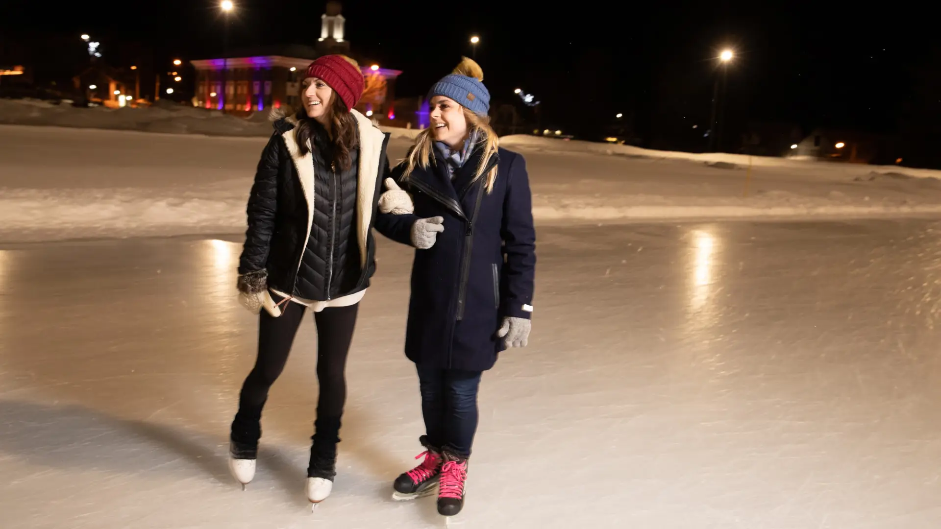 A pair of women in ice skates laughing on an outdoor rink in the evening.