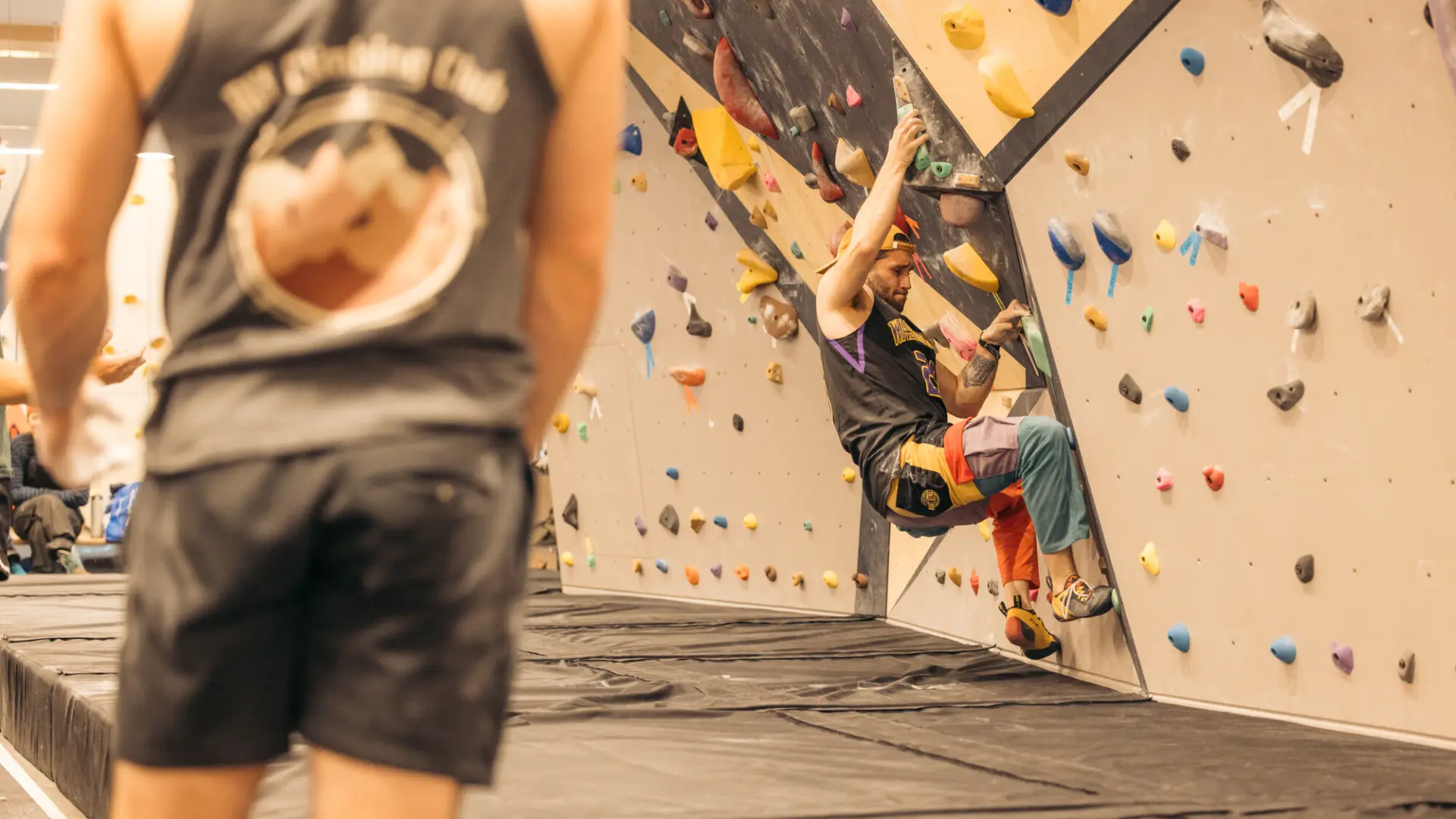 Climber on indoor bouldering wall with person looking on in foreground